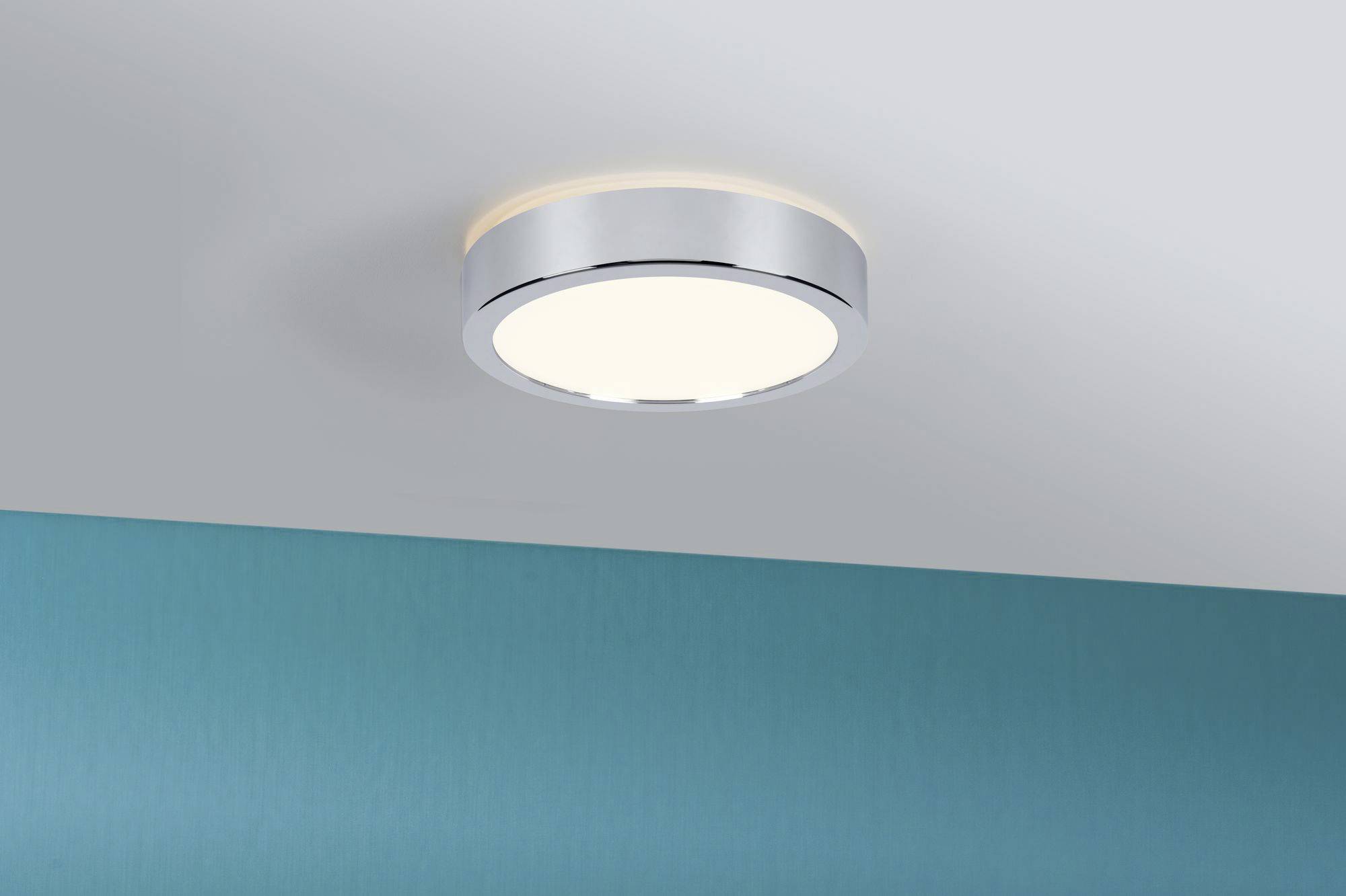Rund loftslampe med sølvfarvet ramme på en hvid loft. Nedenunder ses en turkisfarvet væg.