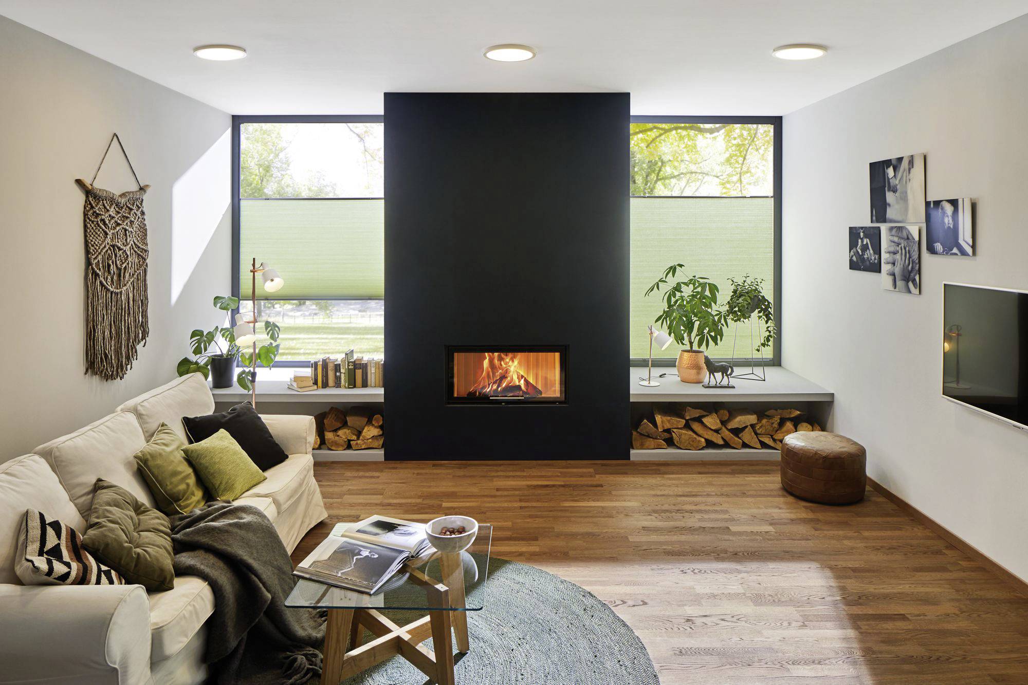 Et moderne opholdsrum med sofa, pejs midt i rummet og store vinduer. Dekorationer og planter skaber en hyggelig atmosfære.
