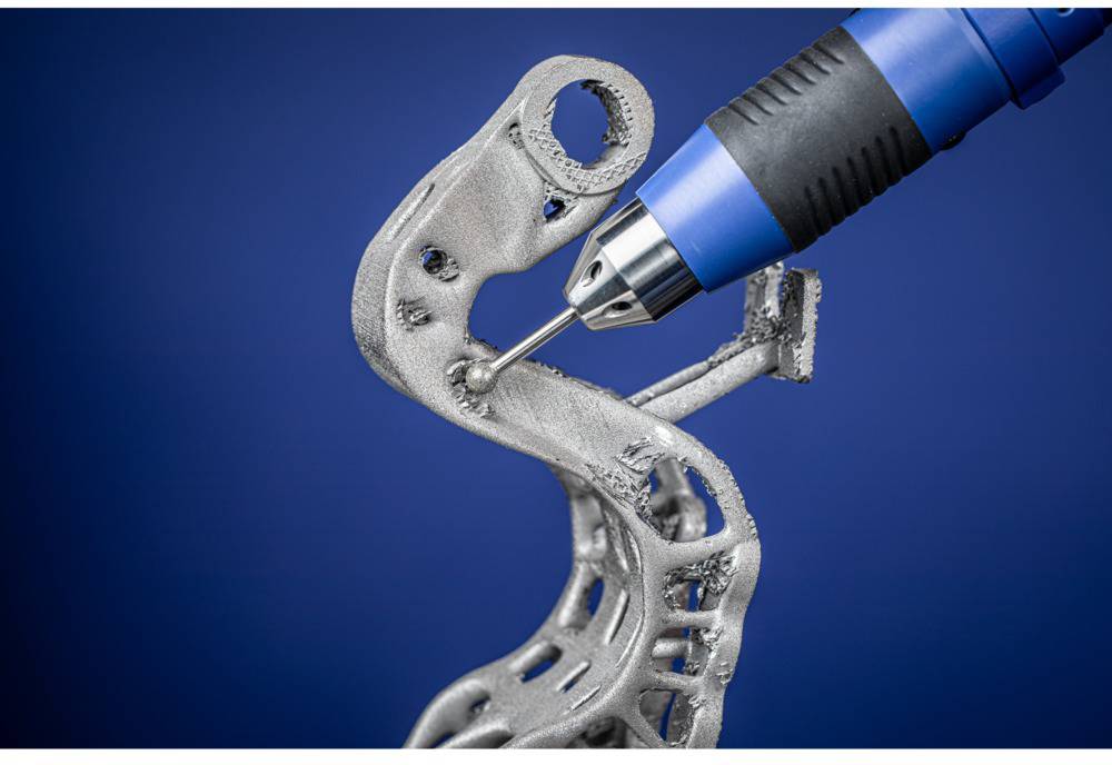 En 3D-printer med et blåt dyse bearbejder en kompleks, metallisk komponent foran en blå baggrund.