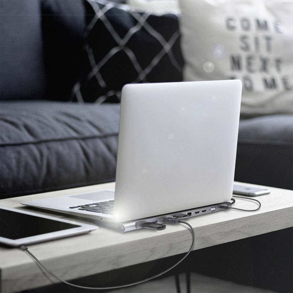 En bærbar computer står på et bord med en USB-hub tilsluttet. I baggrunden ses en sofa med en pude.