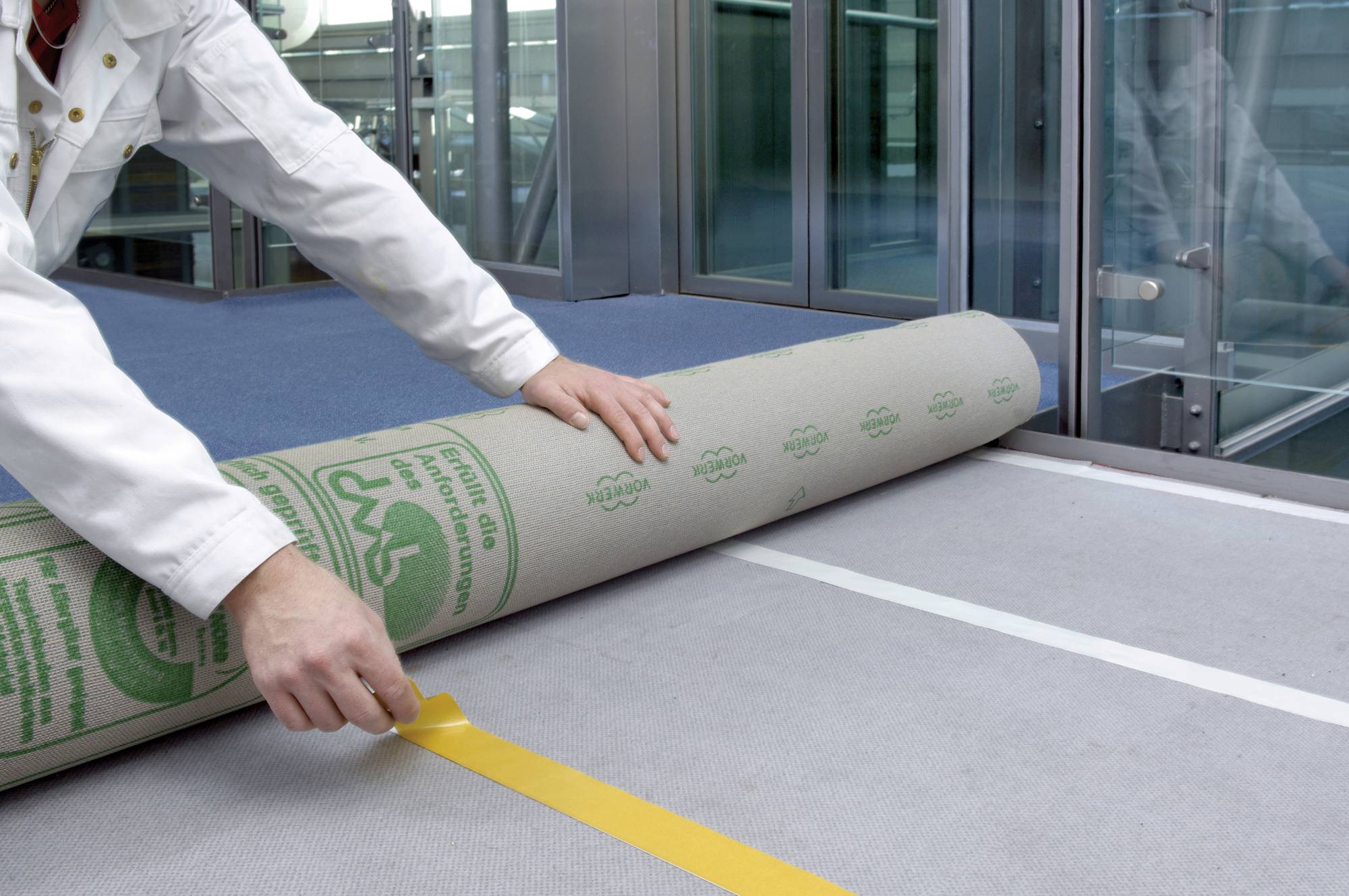 En person lægger en gul klæbende tape på et gråt gulvbelægning i en moderne kontorbygning.