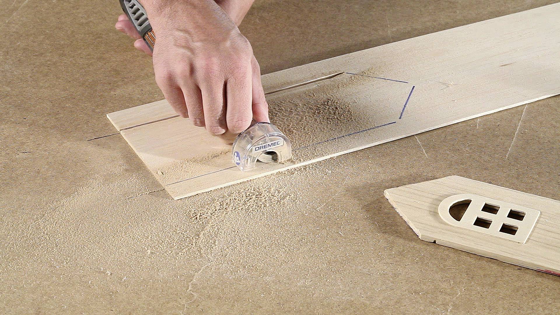 En hånd bruger et elværktøj til at save træ. Savsmuld omgiver arbejdsområdet. Et tilskåret stykke træ ligger ved siden af.