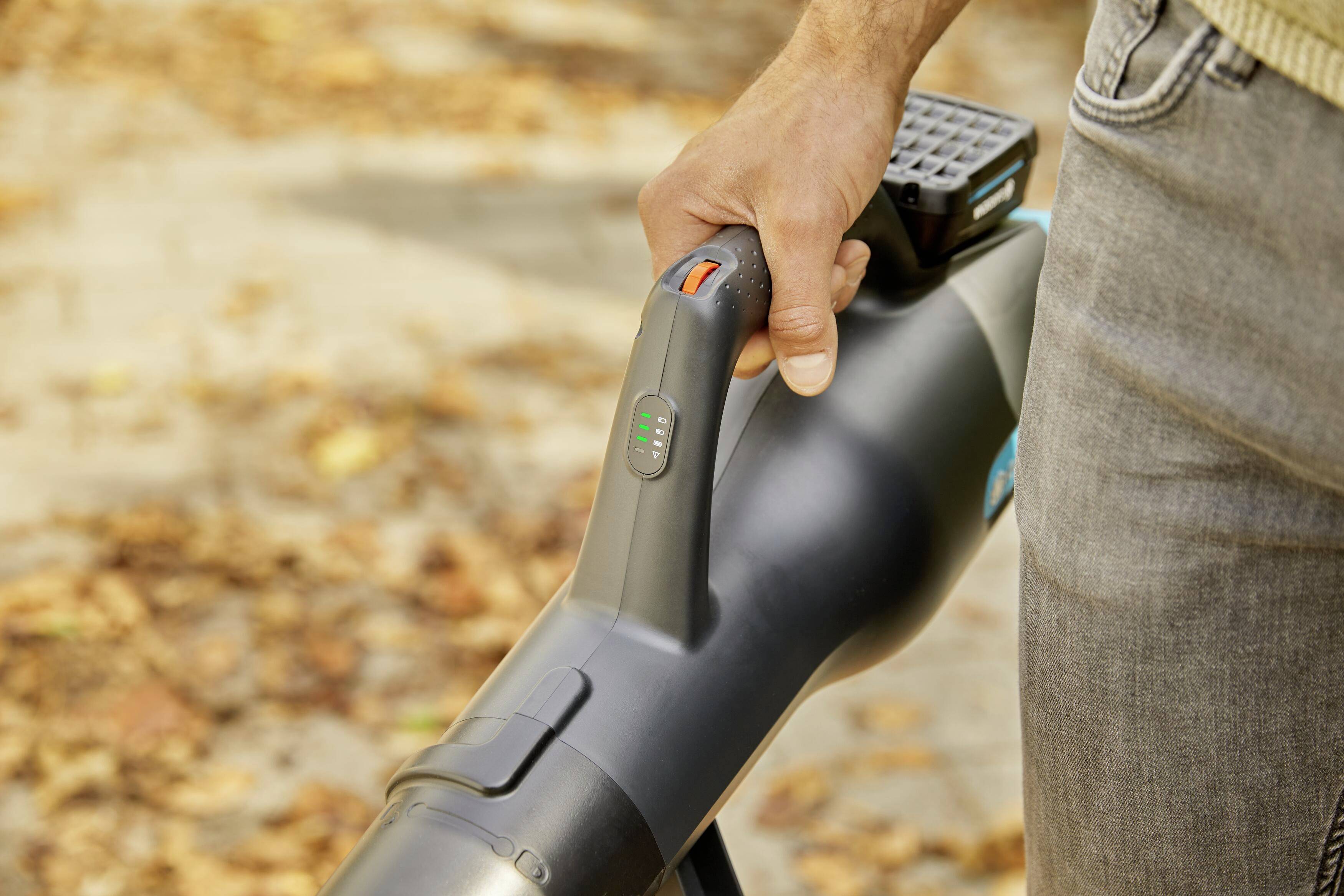 En person holder en løvblæser på en efterårsbelagt gangsti. Enheden er tændt og viser batteristatus.