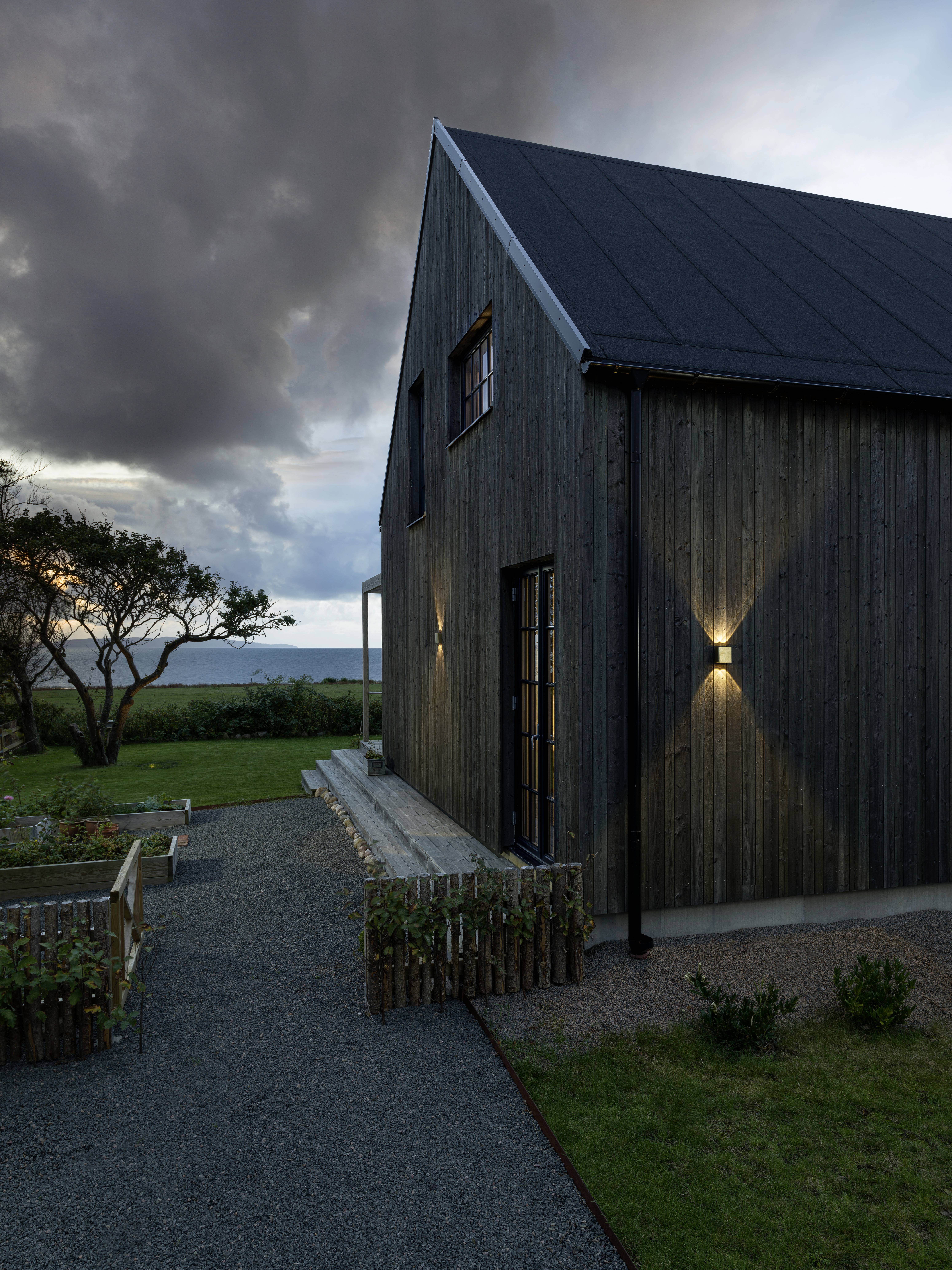 Et moderne træhus ved skumringstid ved kysten, med oplyst facade og have, træer i baggrunden, mørke skyer på himlen.