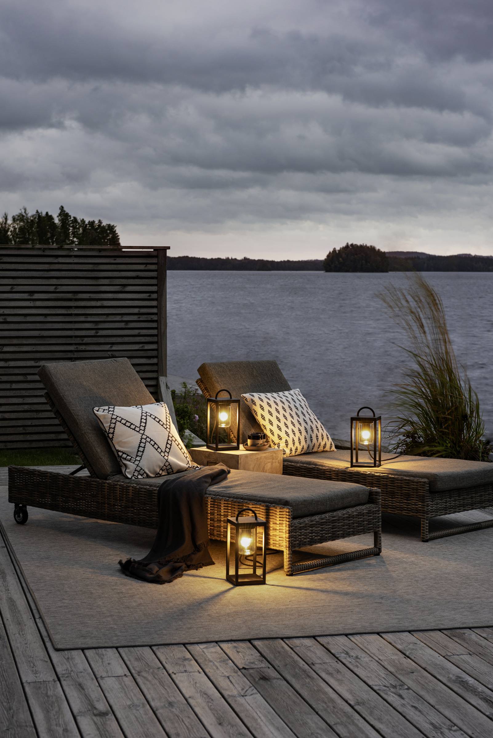 To liggestole står på en terrasse med udsigt over en sø, omgivet af lanterner, der udsender varmt lys. Skyet himmel i baggrunden.