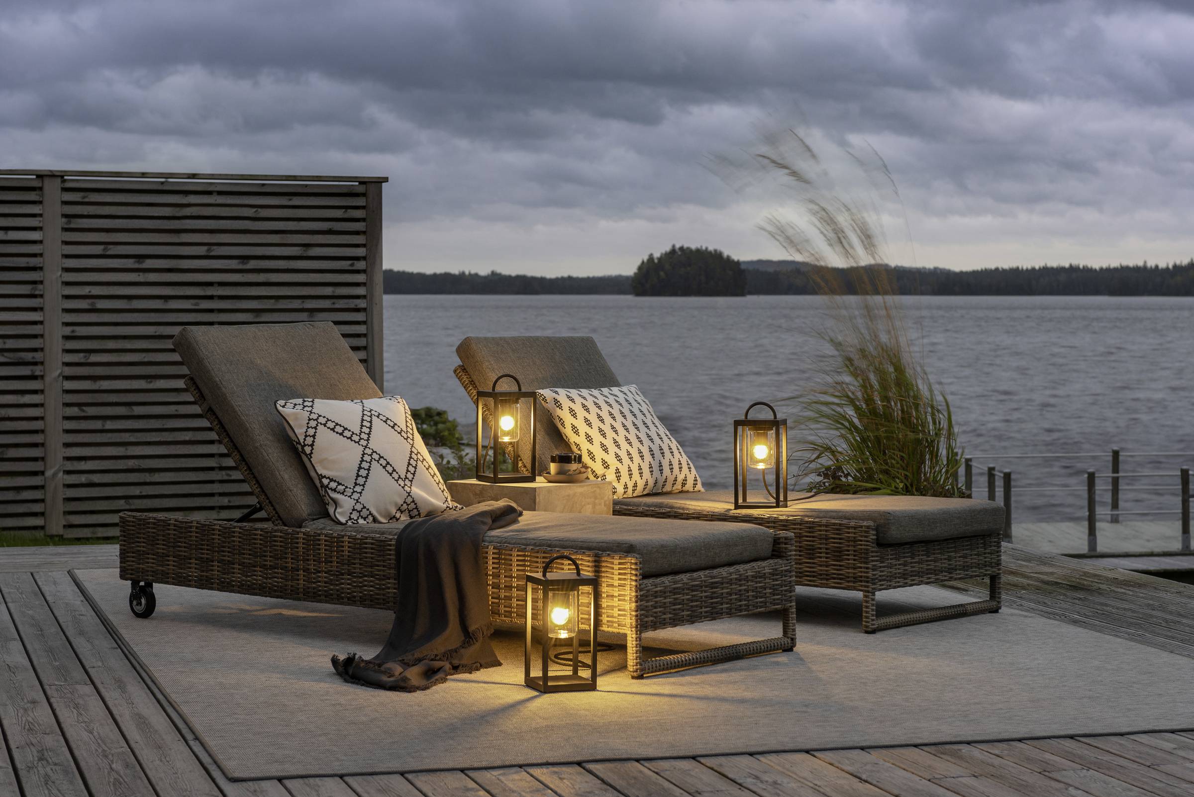 En terrasse med udsigt over en sø under en overskyet himmel. To liggestole med lanterner, tæpper og puder skaber en hyggelig atmosfære.