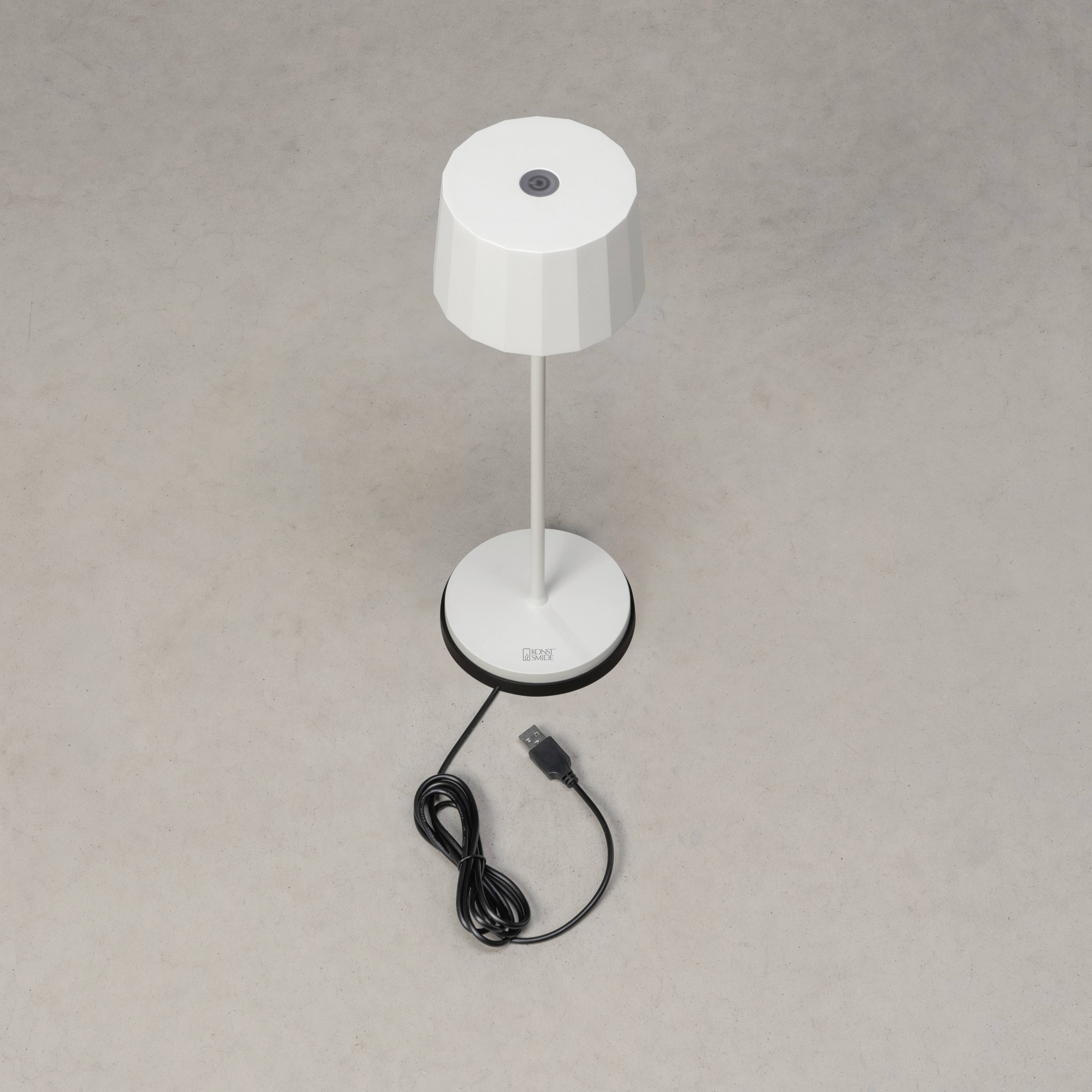 En hvid bordlampe med rund skærm står på en lys baggrund. Kablet med USB-stik er viklet rundt om lampens sokkel.