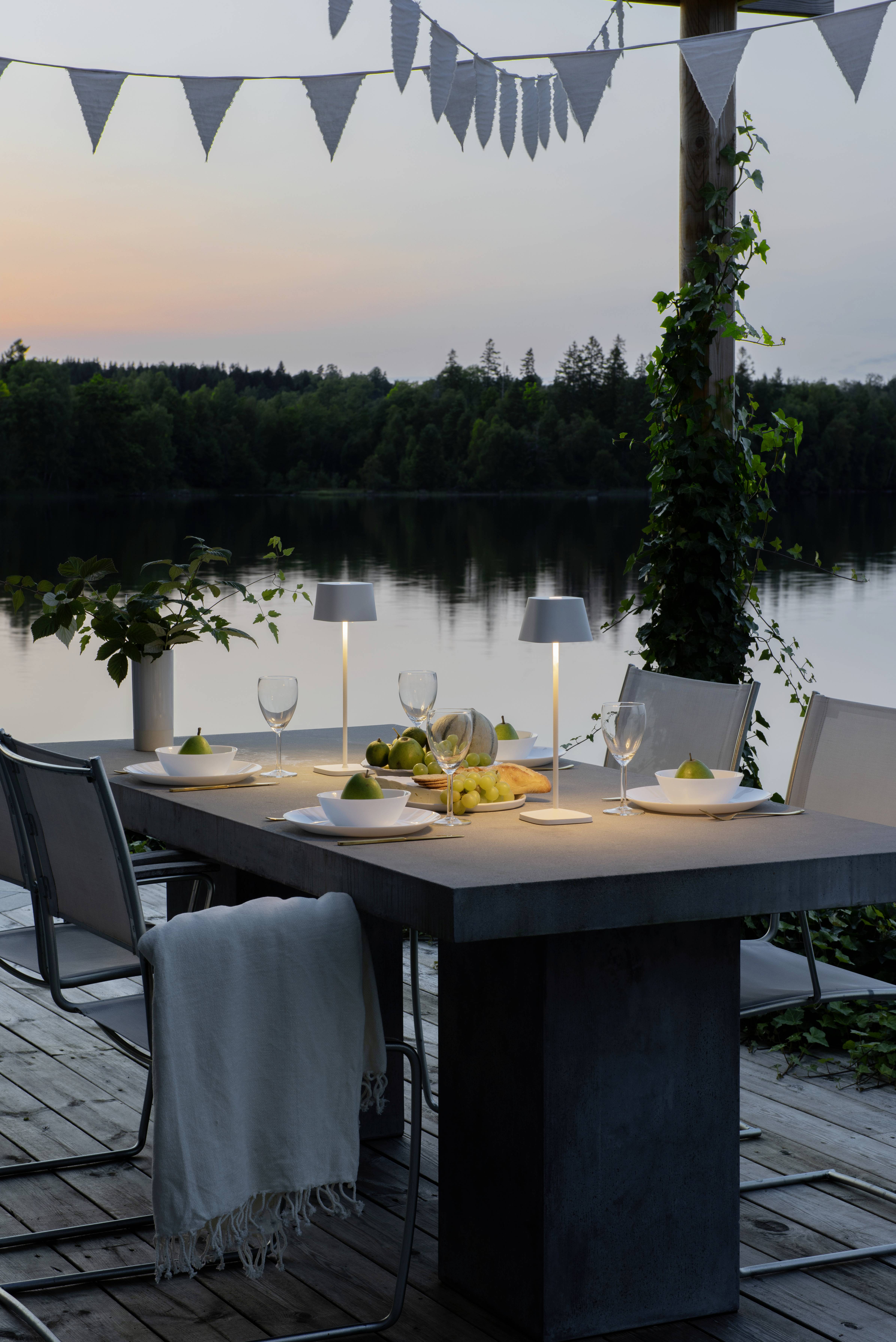 Et dækket bord ved solnedgang, udendørs på en træterrasse ved søen. Lamper oplyser glas, tallerkener og pærer.