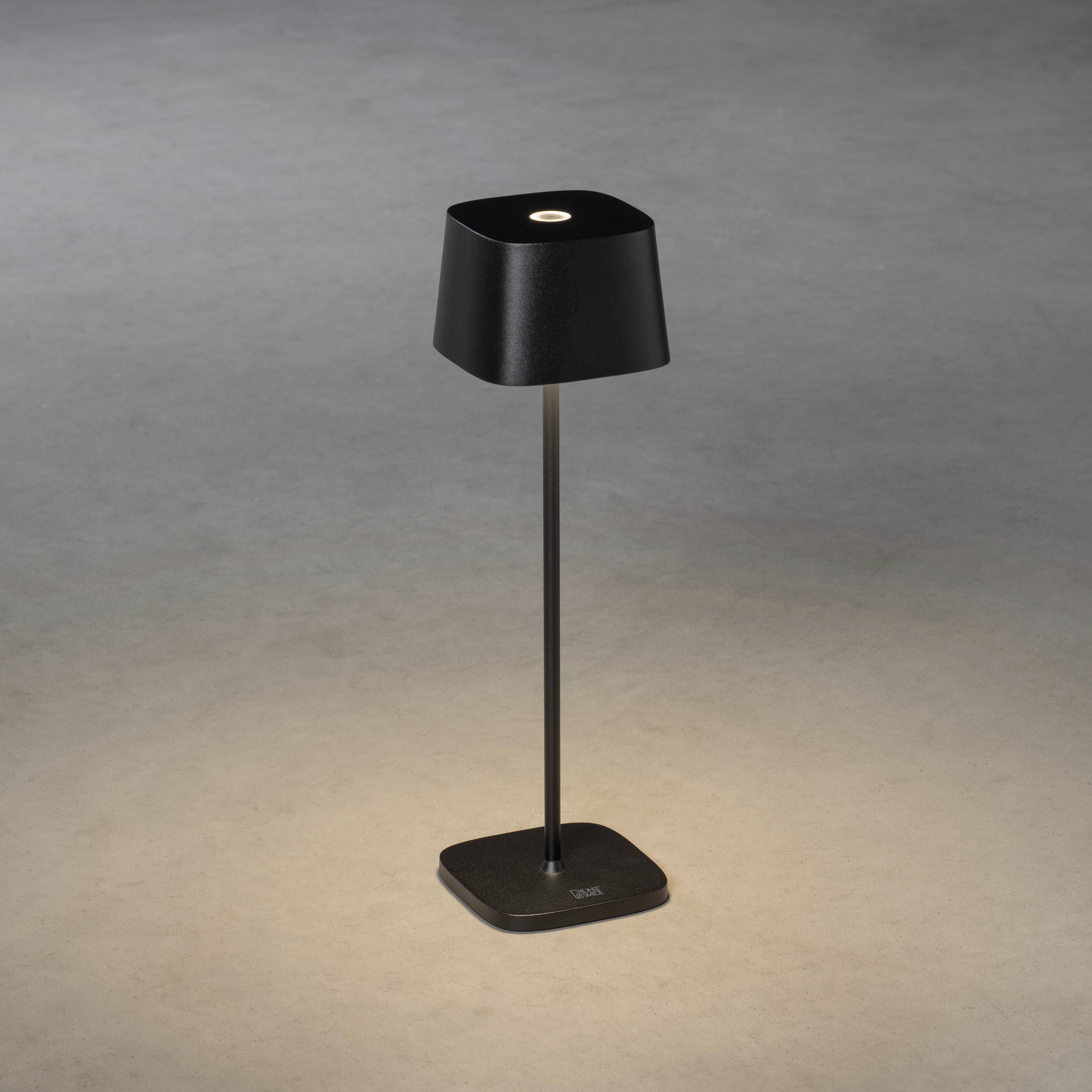 Sort bordlampe med firkantet skærm på en slank fod, som udsender varmt lys ned på det grå gulv.