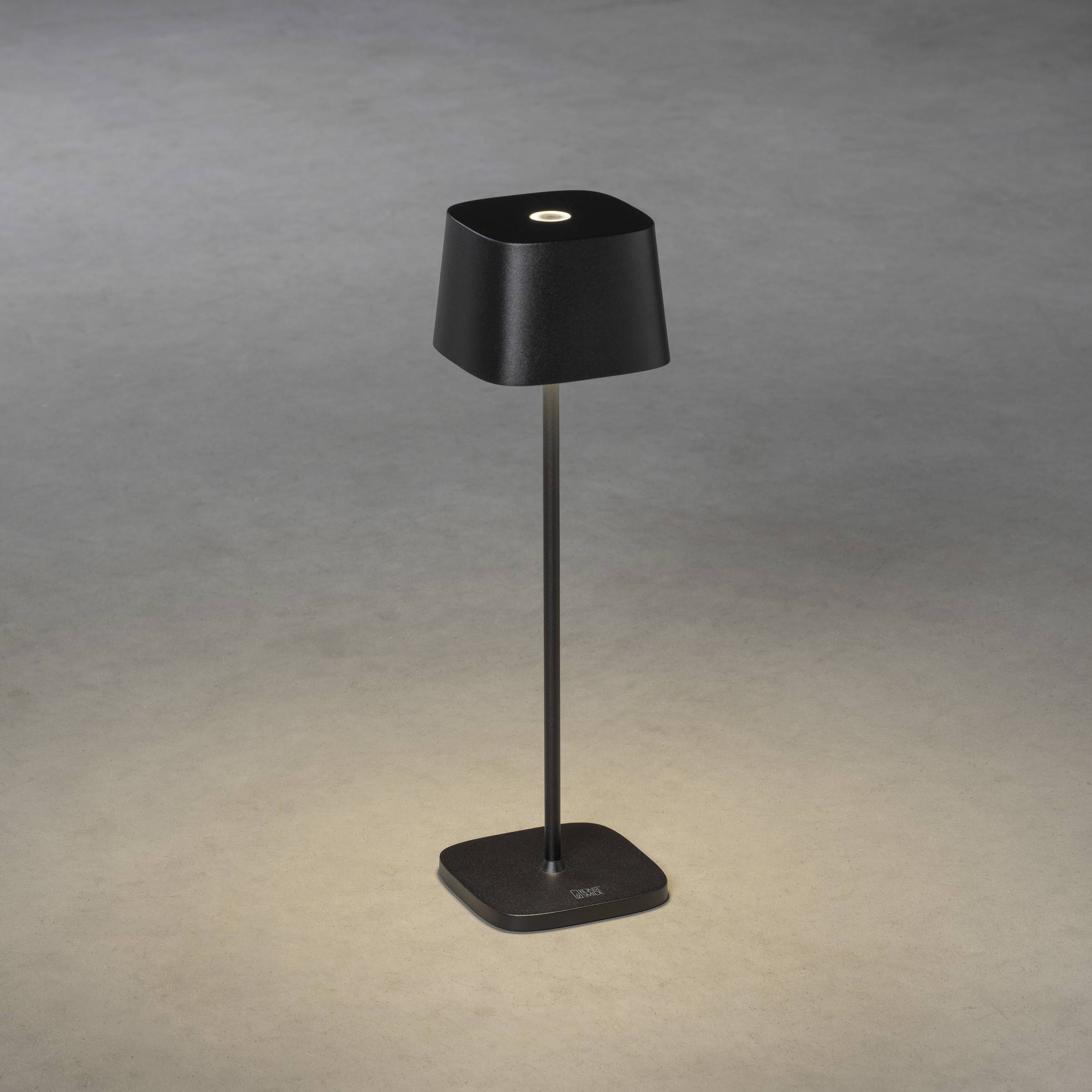 En slank sort bordlampe med en firkantet skærm belyser gulvet punktvist i et skyggefuldt belyst rum.