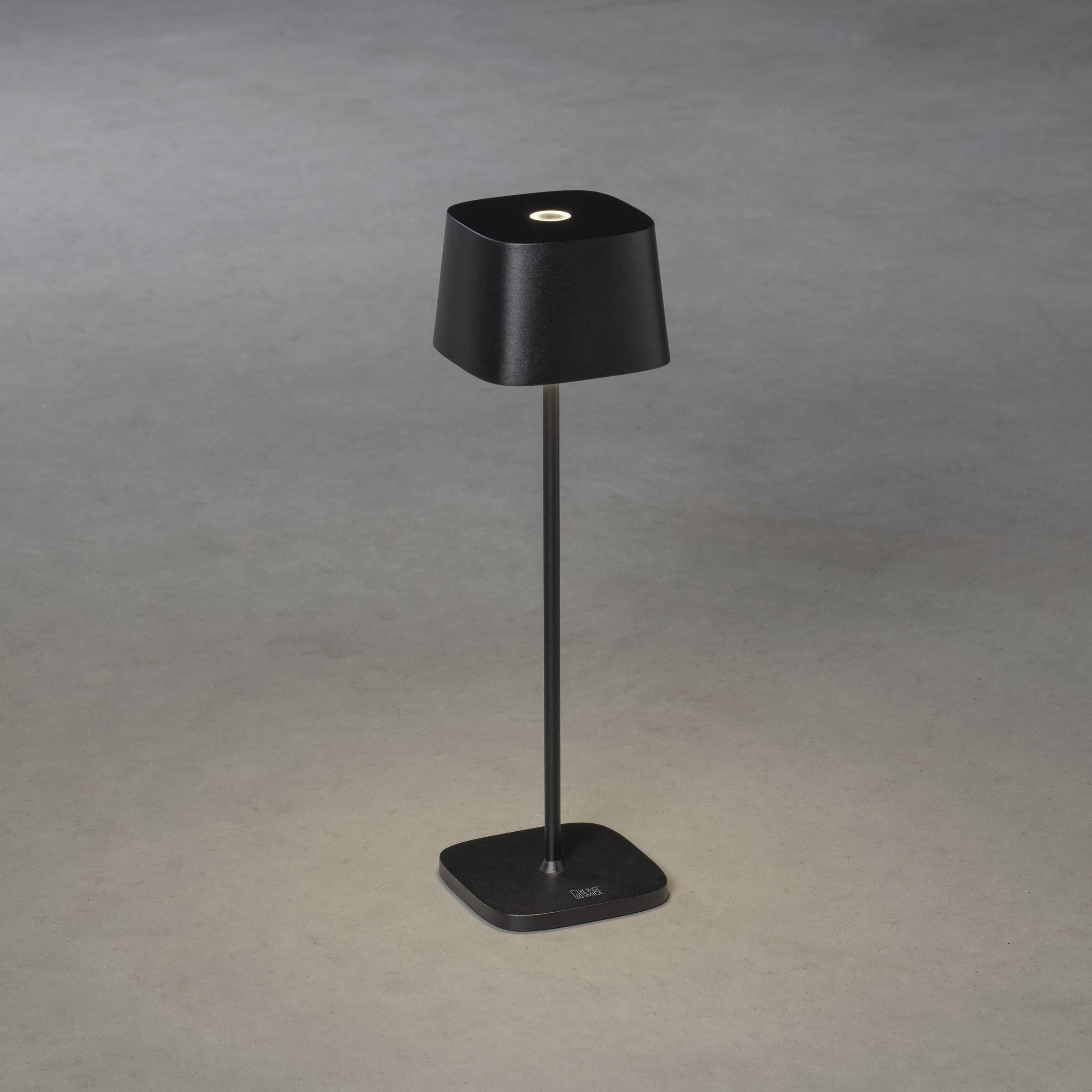 En sort bordlampe med en firkantet skærm og fod står på et gråt underlag og lyser nedad.