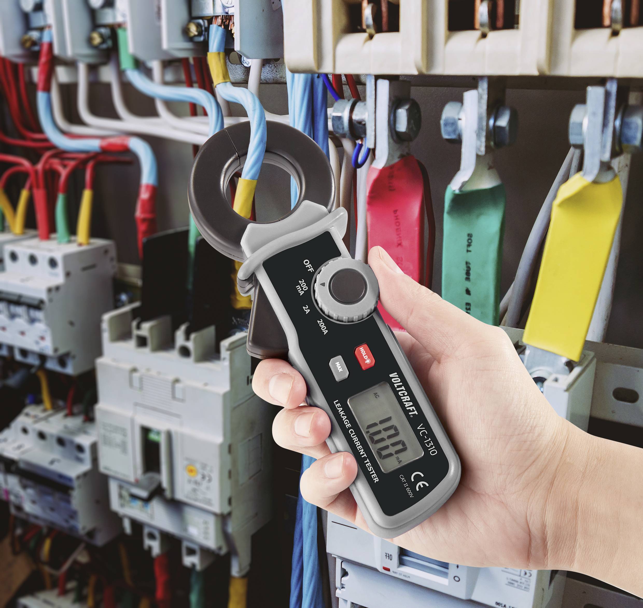 En hånd holder et digitalt multimeter med strømtang foran et åbent el-skab med farvede kabler og sikringer.