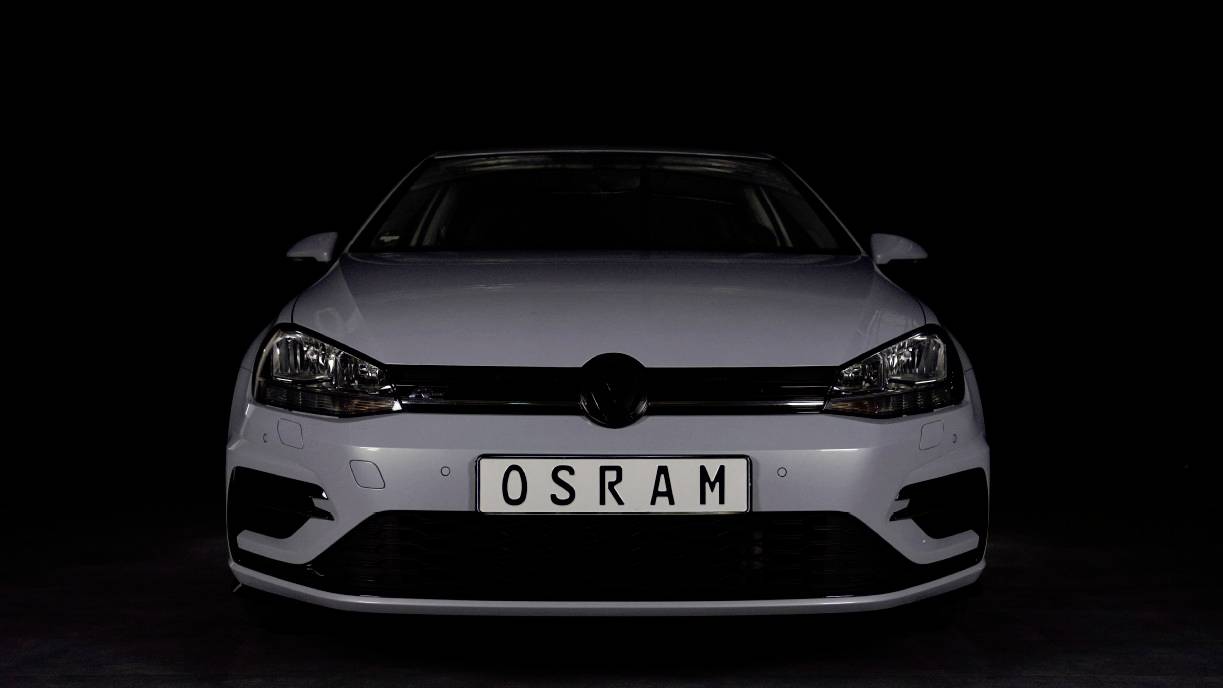 En hvid bil set forfra med et nummerplade, hvorpå der står 'OSRAM', foran en sort baggrund.