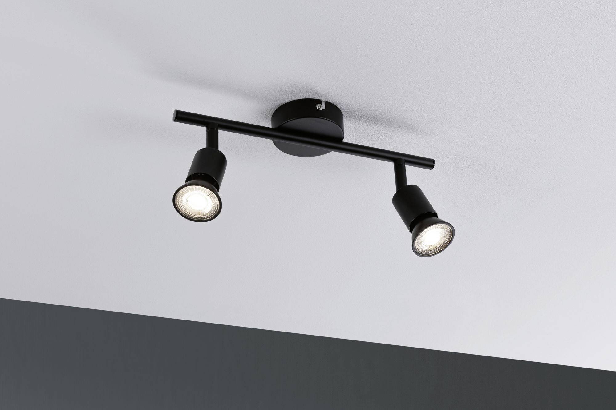 Sort loftslampe med to justerbare projektører, monteret på en hvid loft. Oplyser et moderne rum.