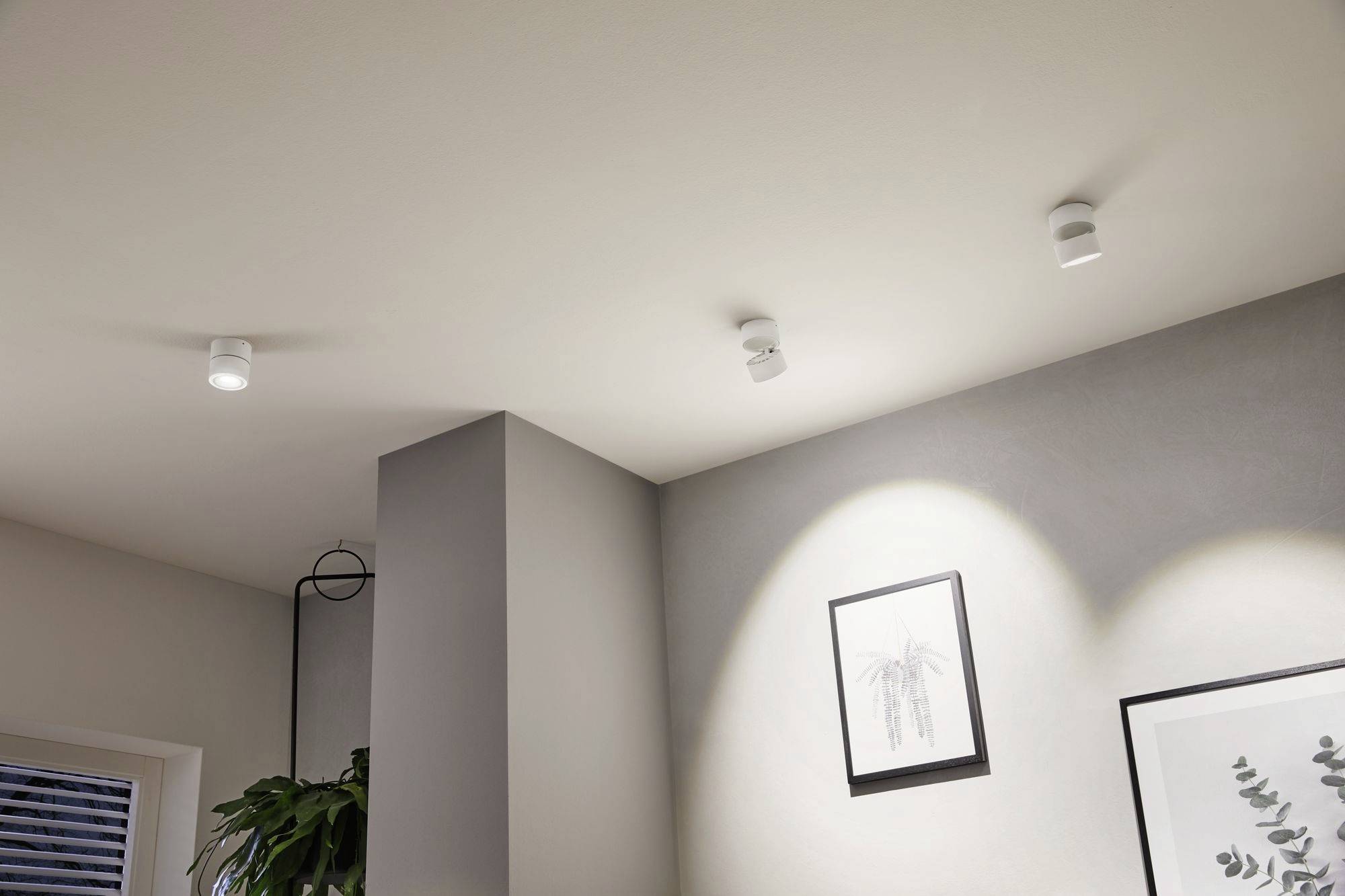 Tre hvide loftslamper oplyser et moderne stue. En plante står i hjørnet, og ved siden af hænger to indrammet billeder.