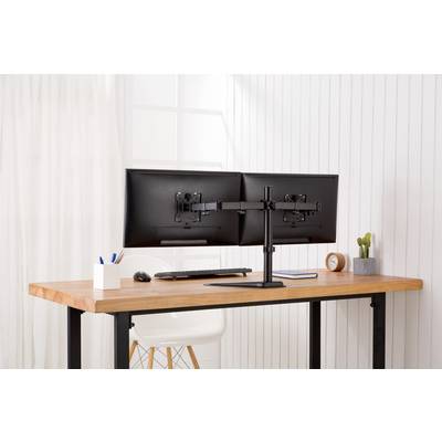 To schwarze computerskærme på et træskrivebord, ved siden af en penneholder, en mus og et ur i et hvidt rum.