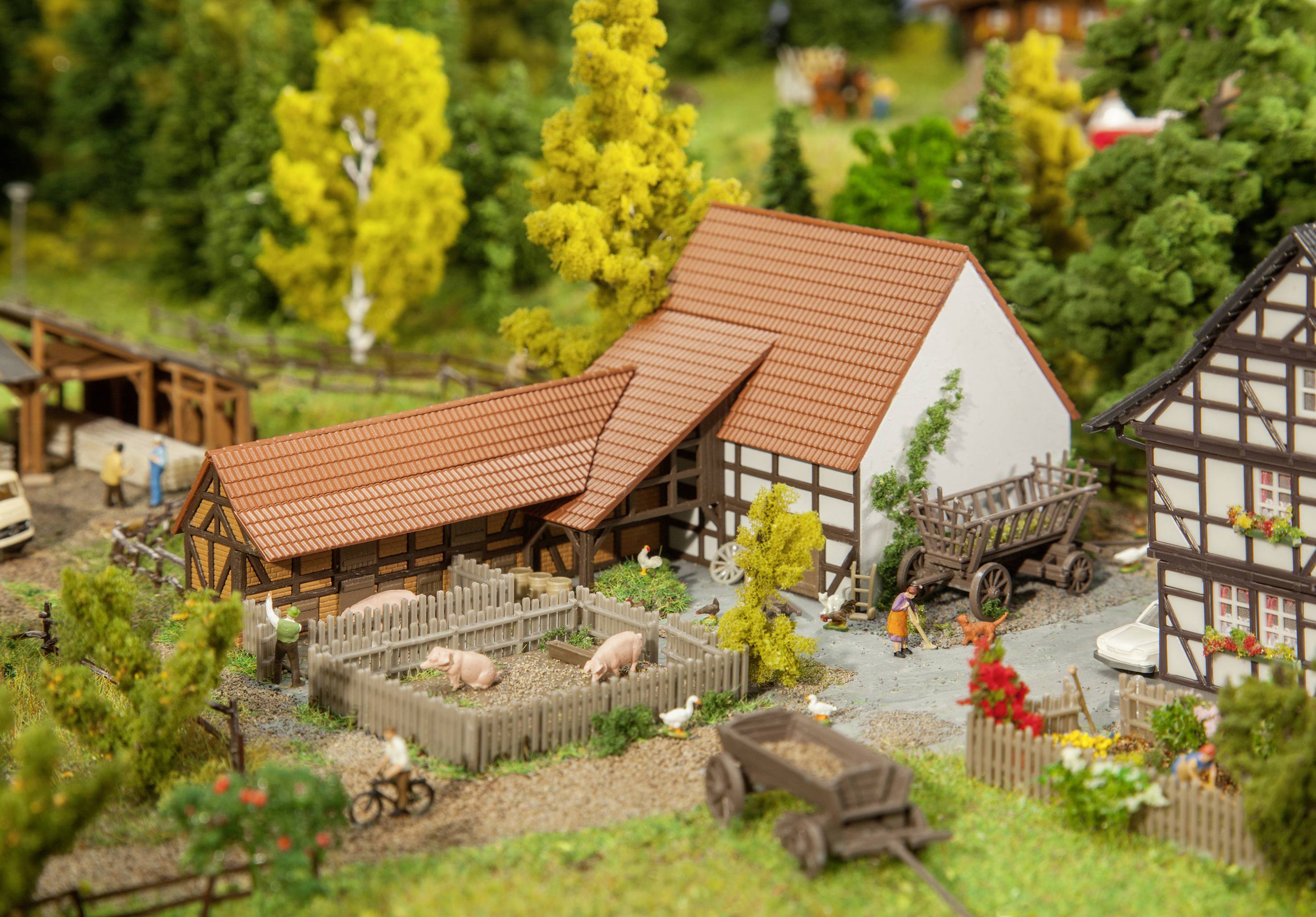 En miniature bondegård med bindingsværkshuse, grise i indhegningen og en trækvogn. Omgivet af træer og figurer på en grusvej.