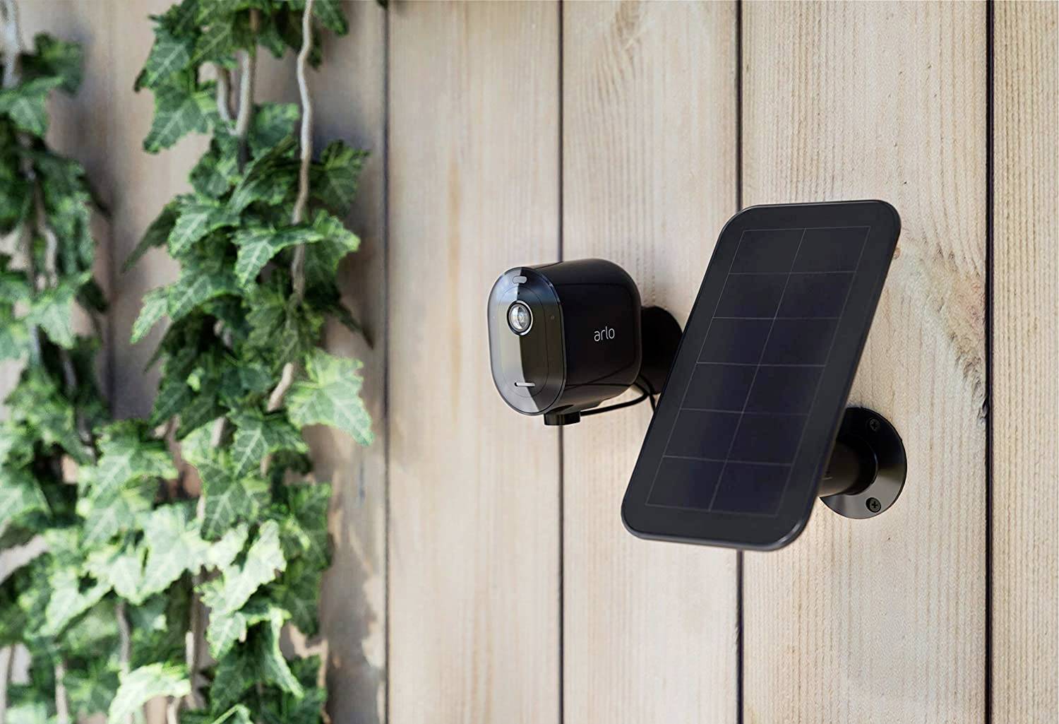 Sikkerhedskamera med solarpanel, monteret på en træmur ved siden af en plante.