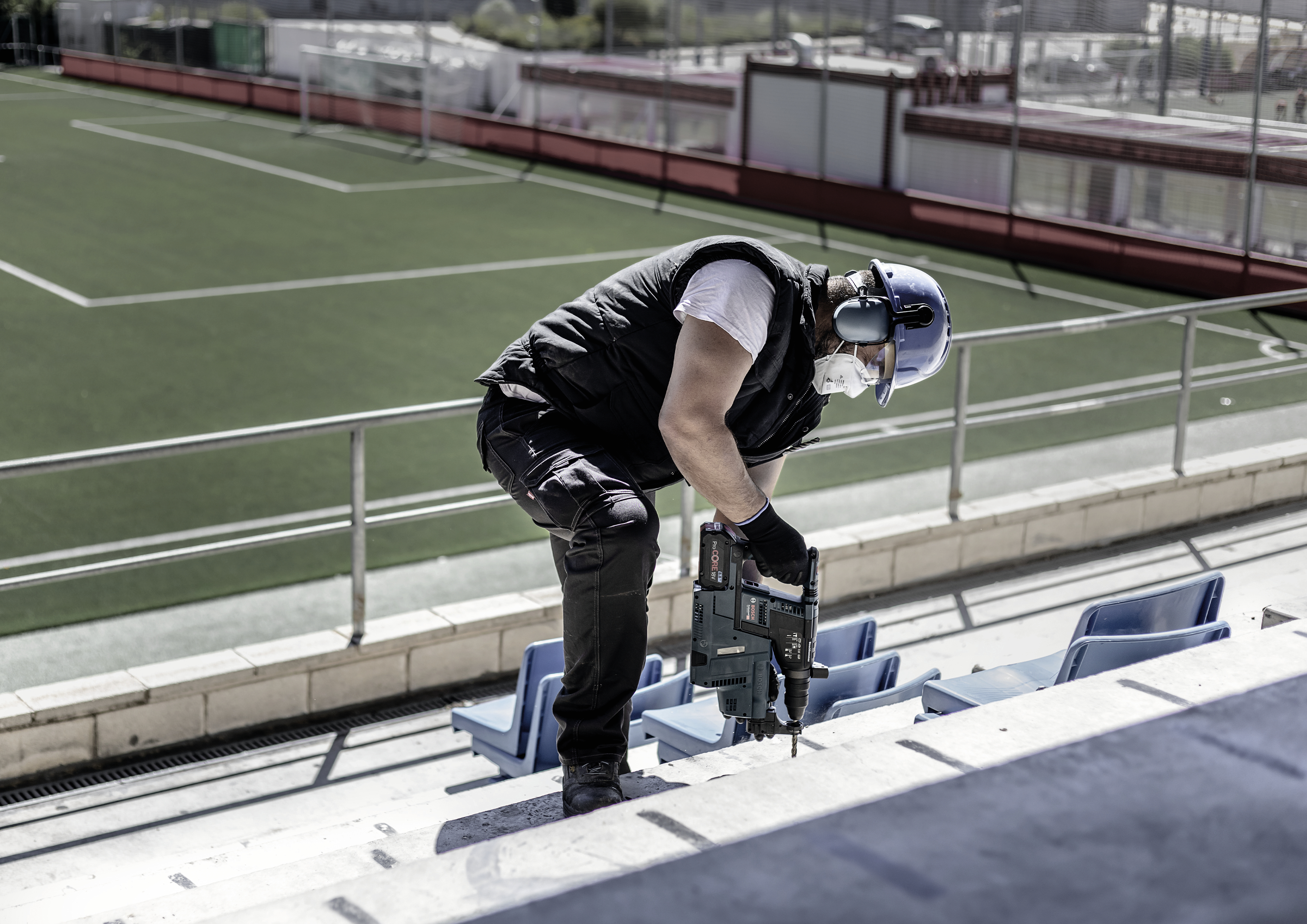 En arbejder med sikkerhedshjelm og høreværn udfører bygningsarbejde på trinene i et tomt stadion.
