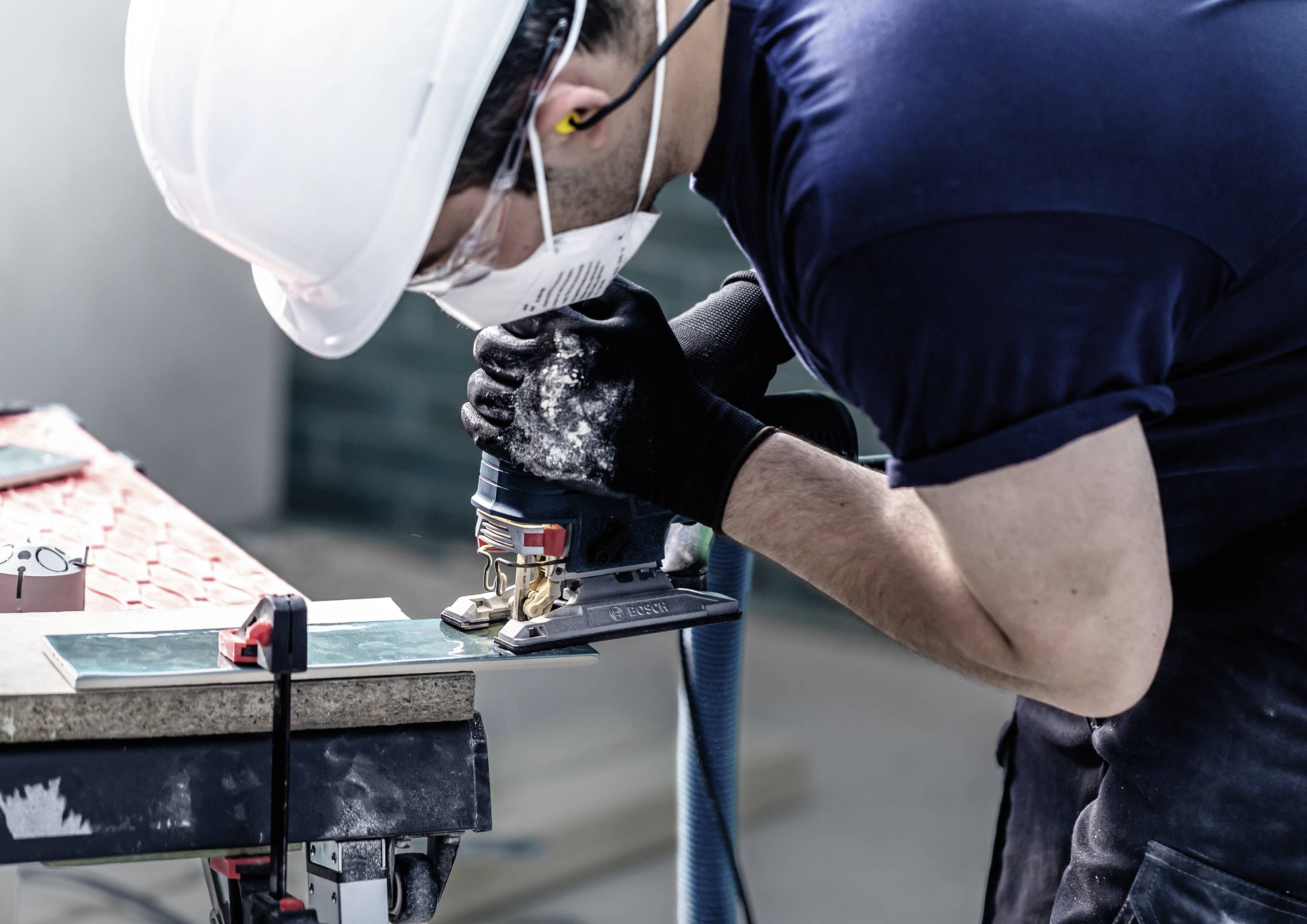 En person med beskyttelseshjelm og maske arbejder med et elektrisk værktøj på et stykke træ i et værkstedsmiljø.