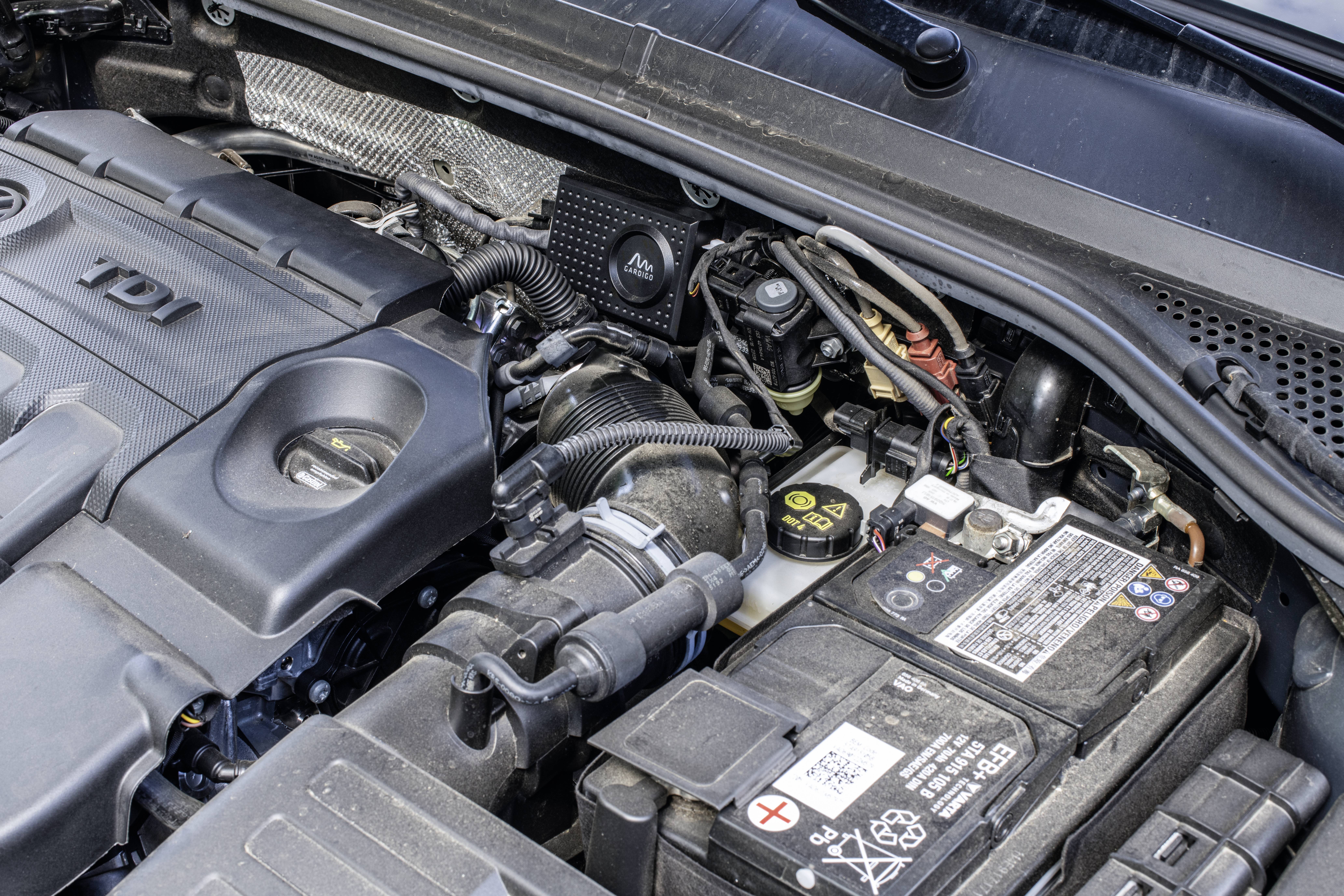 Motorrummet i et køretøj, viser dele som bilbatteri og motorafdækning. Ledningsnet og slanger er tydeligt synlige.