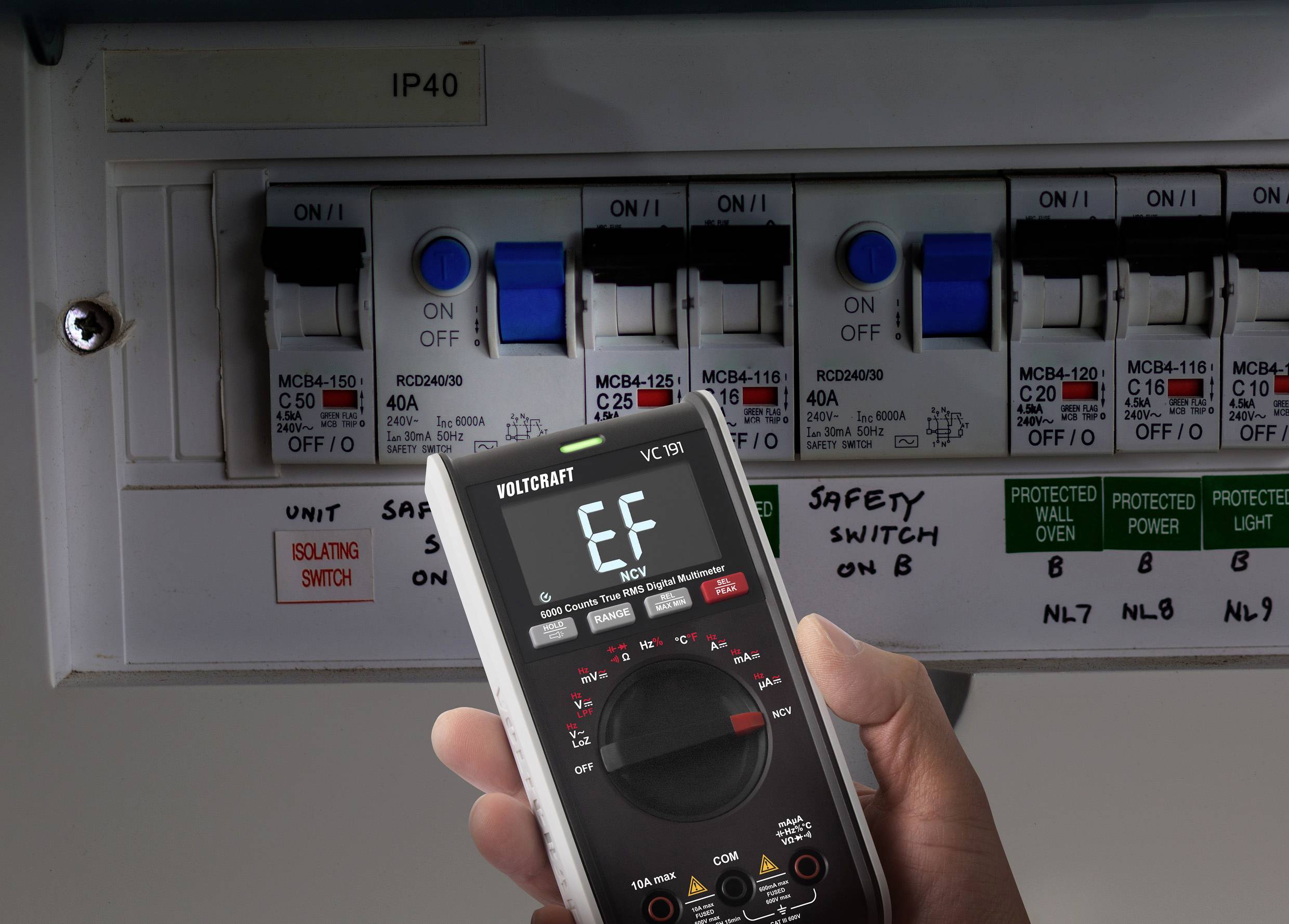 En person holder et digitalt multimeter, der viser en måleværdi, foran en el-tavle med flere sikringskontakter i en bygning.