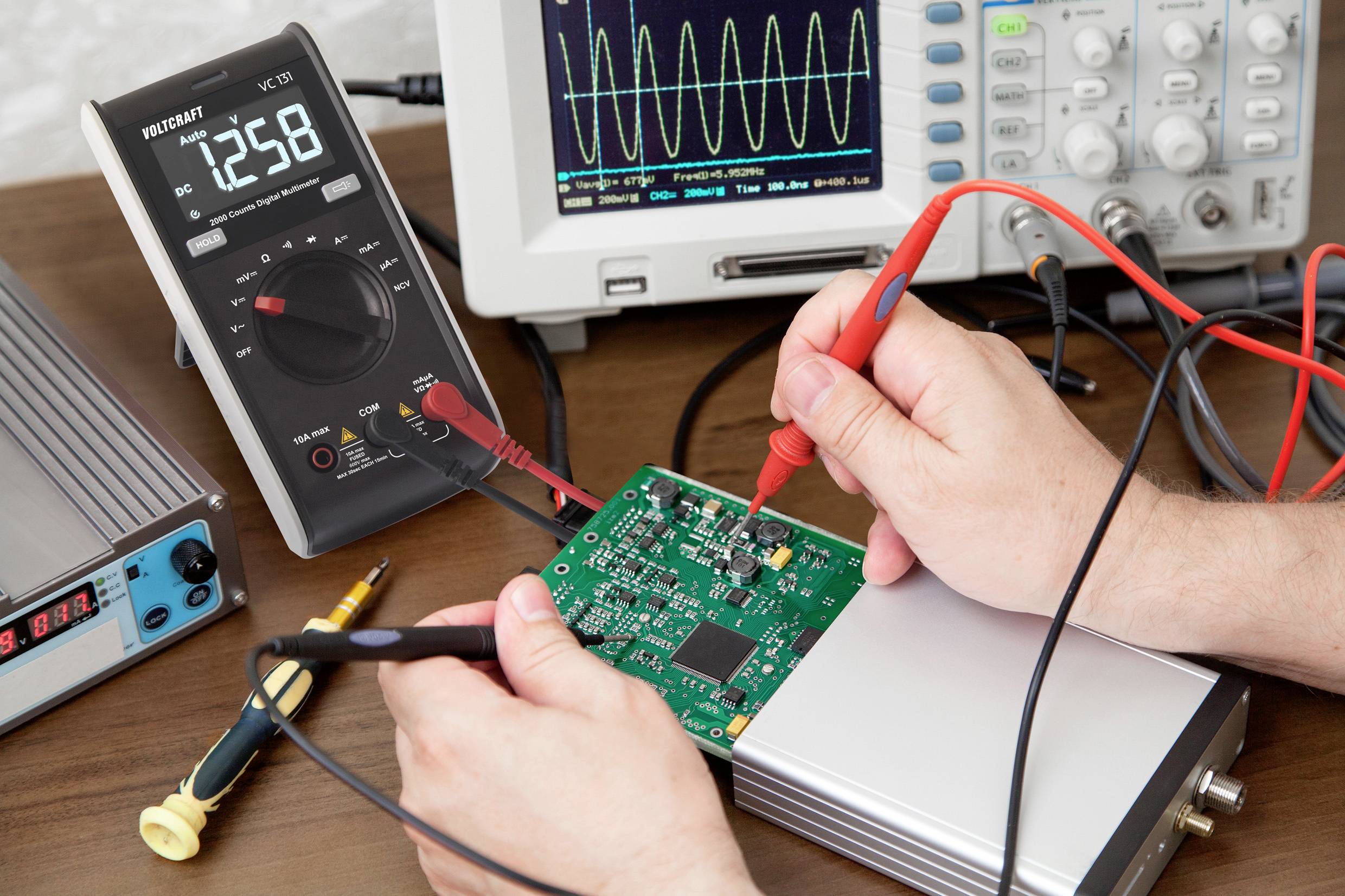 En person tester et grønt elektronisk kredsløbskort med et multimeter, der ligger på et bord. Oscilloskopskærm i baggrunden.