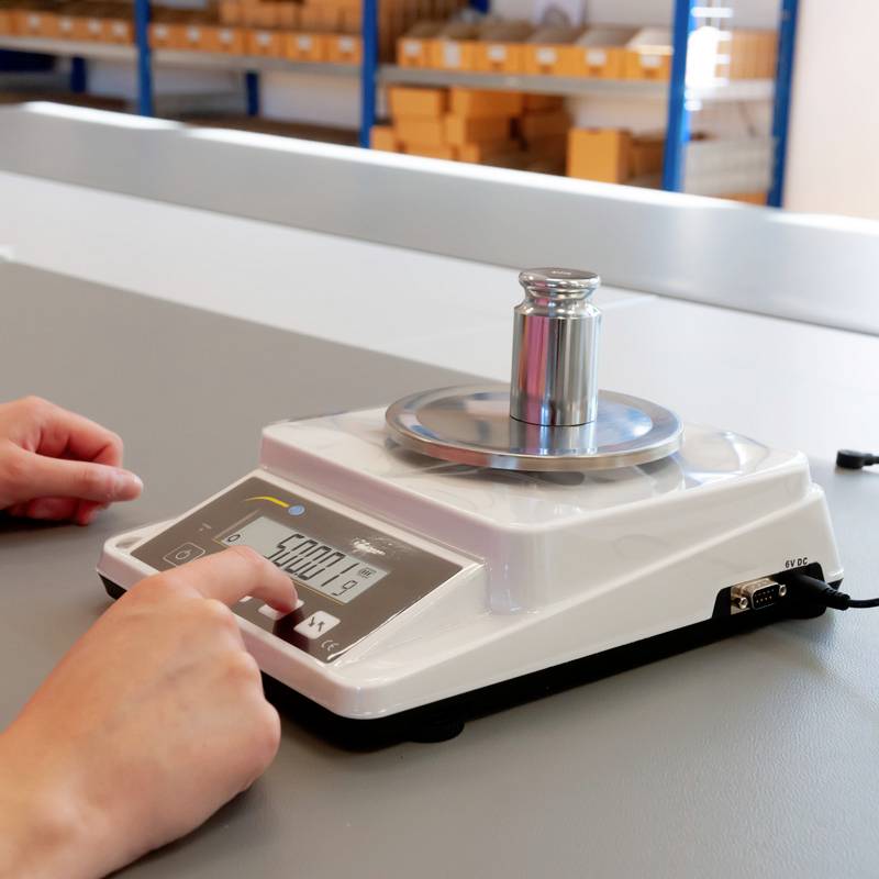 Digital vægt med vægt på platformen viser '5000,19' gram. To hænder er synlige; fokus på måleapparatet.