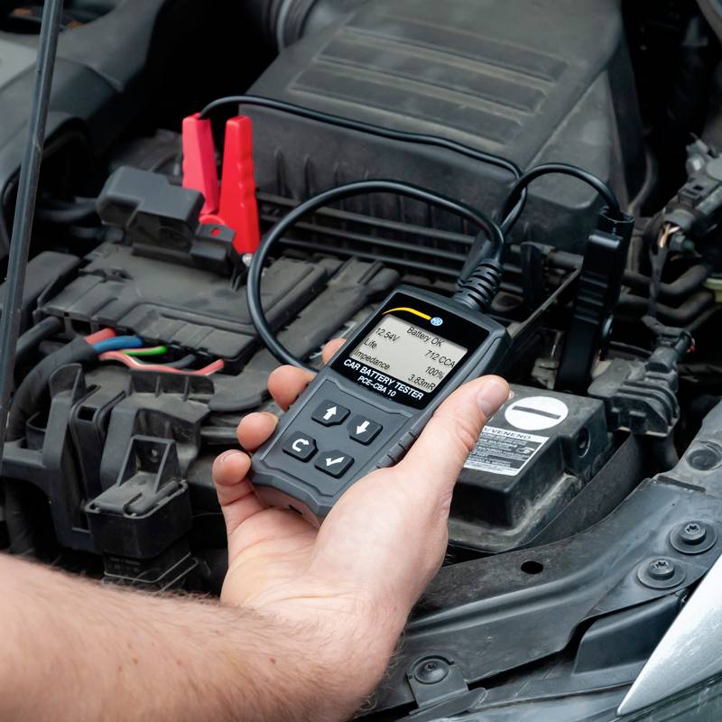 Et håndholdt apparat tester bilbatteriet i en motorrum. En mand holder enheden, som viser data om batteriets tilstand.