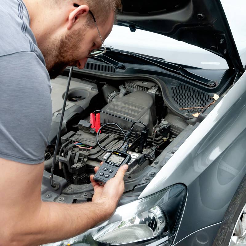 En person tester batteriet i en åben bil med et multimeter.