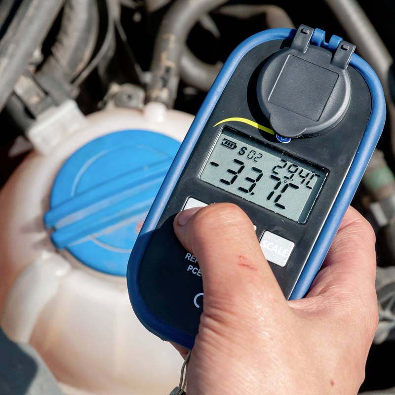 En hånd holder en digital kølervæske-måler, der viser '-33,7°C'. I baggrunden ses en kølervæskebeholder.