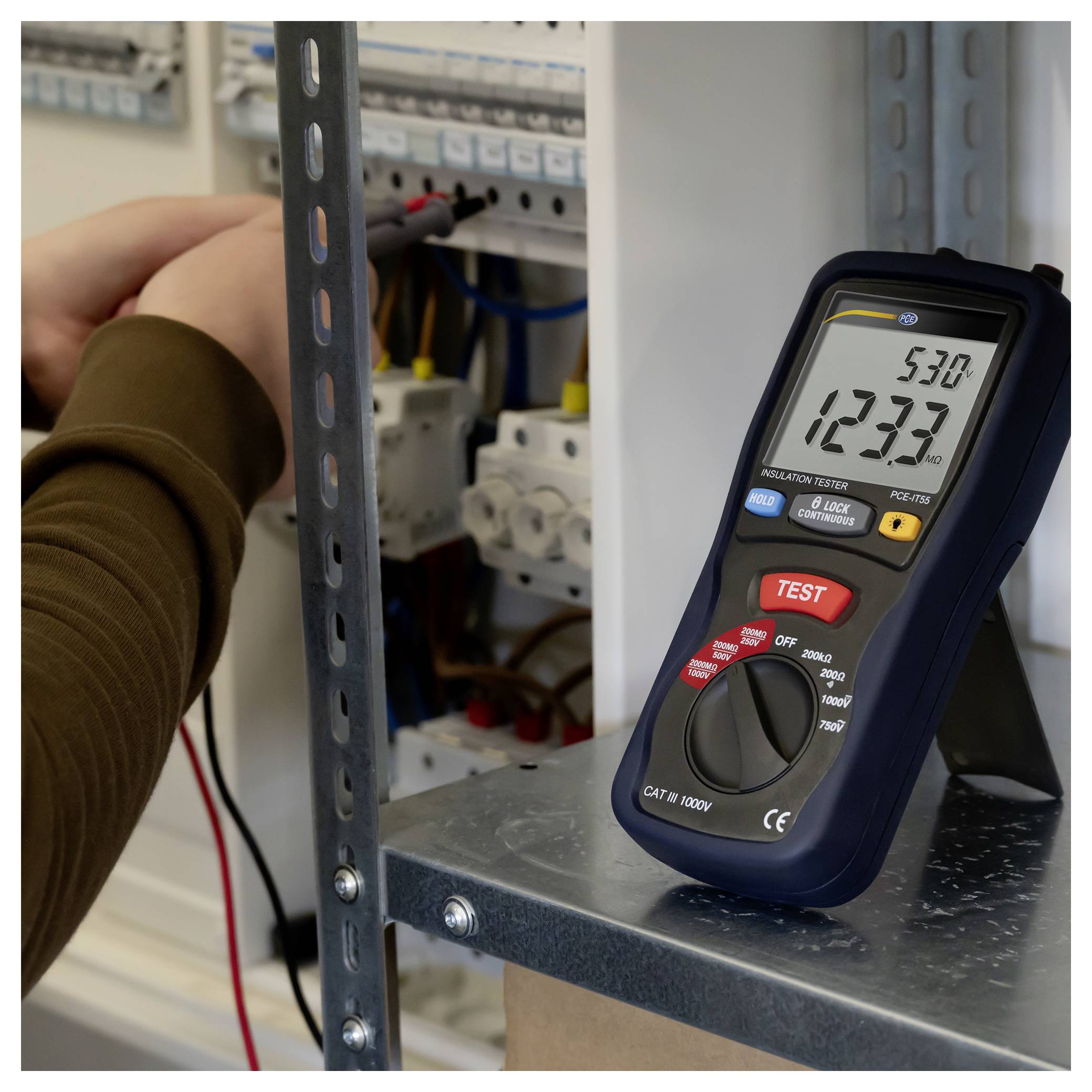 Håndholdt elektrisk tester viser 123,3 volt AC og 53,0 Hz ved siden af en person, der arbejder inden i et elektrisk panel.