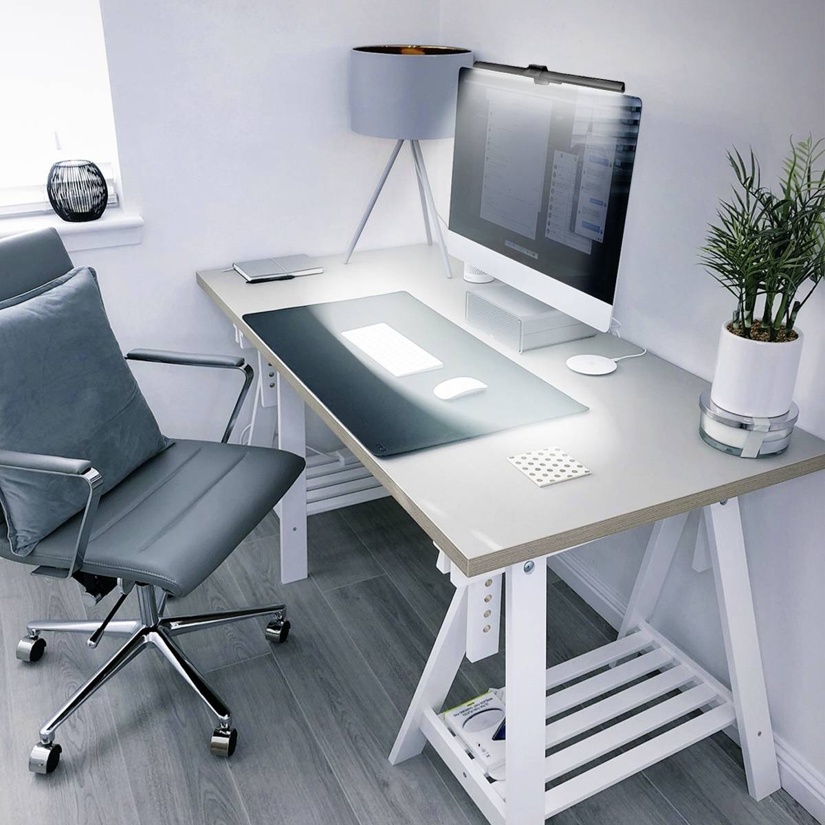 Moderne arbejdsplads med grå kontorstol, hvidt skrivebord, computer, lampe og plante ved siden af et vindue i et hjørnekontorrum.