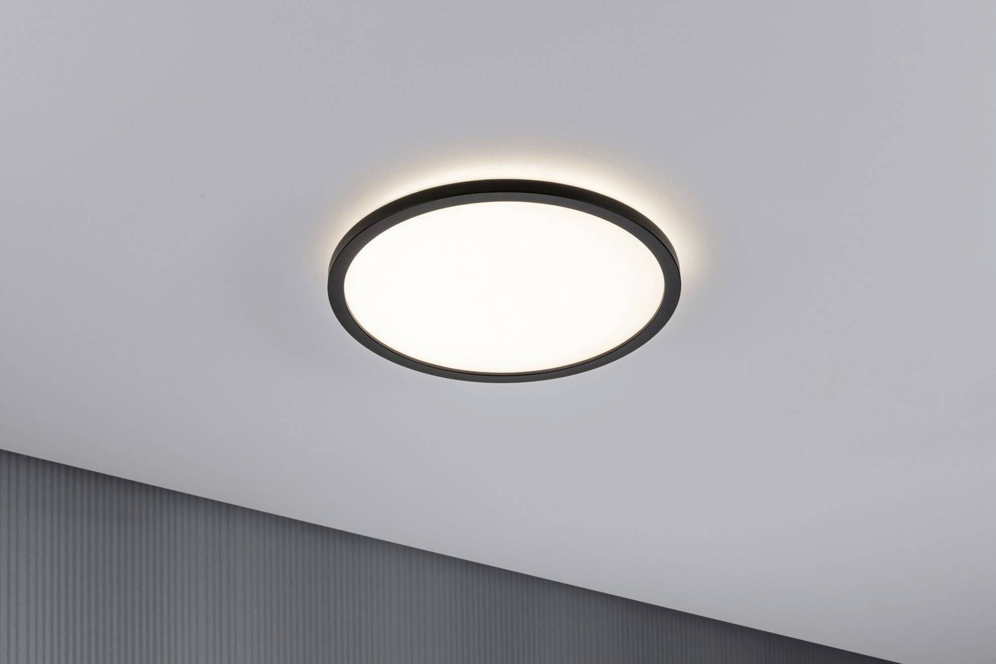 Rund loftlampe med sort ramme på en hvid loftvæg. Lampen er tændt og udsender et klart, kraftigt lys.