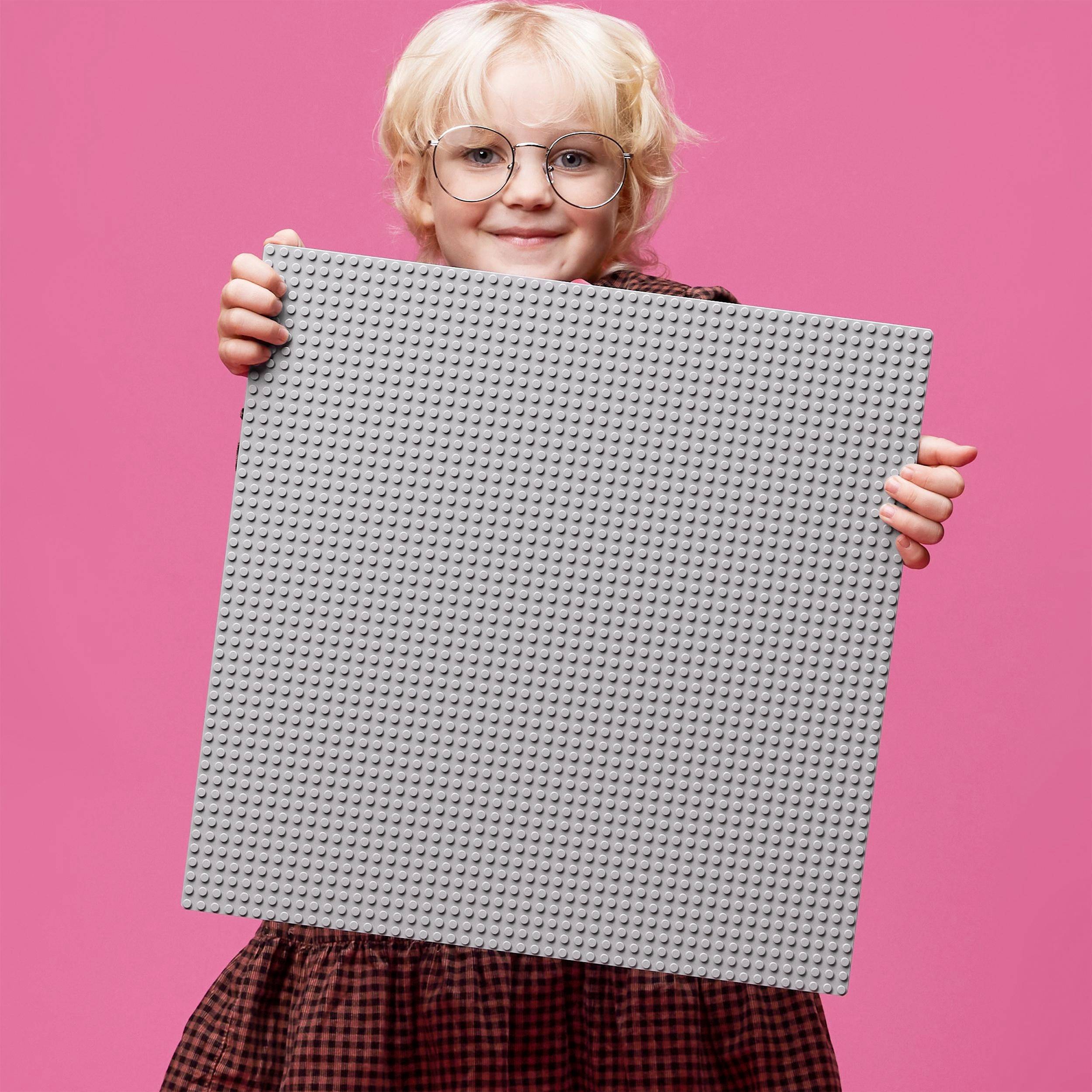 Et barn med lyst hår og briller holder en stor, grå LEGO-byggeplade foran en lyserød baggrund.