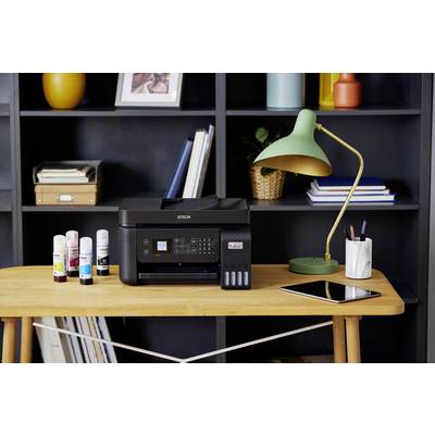 Et skrivebord med en multifunktionsprinter, ved siden af blækflasker. I baggrunden et bogskab, på bordet en lampe og en tablet.