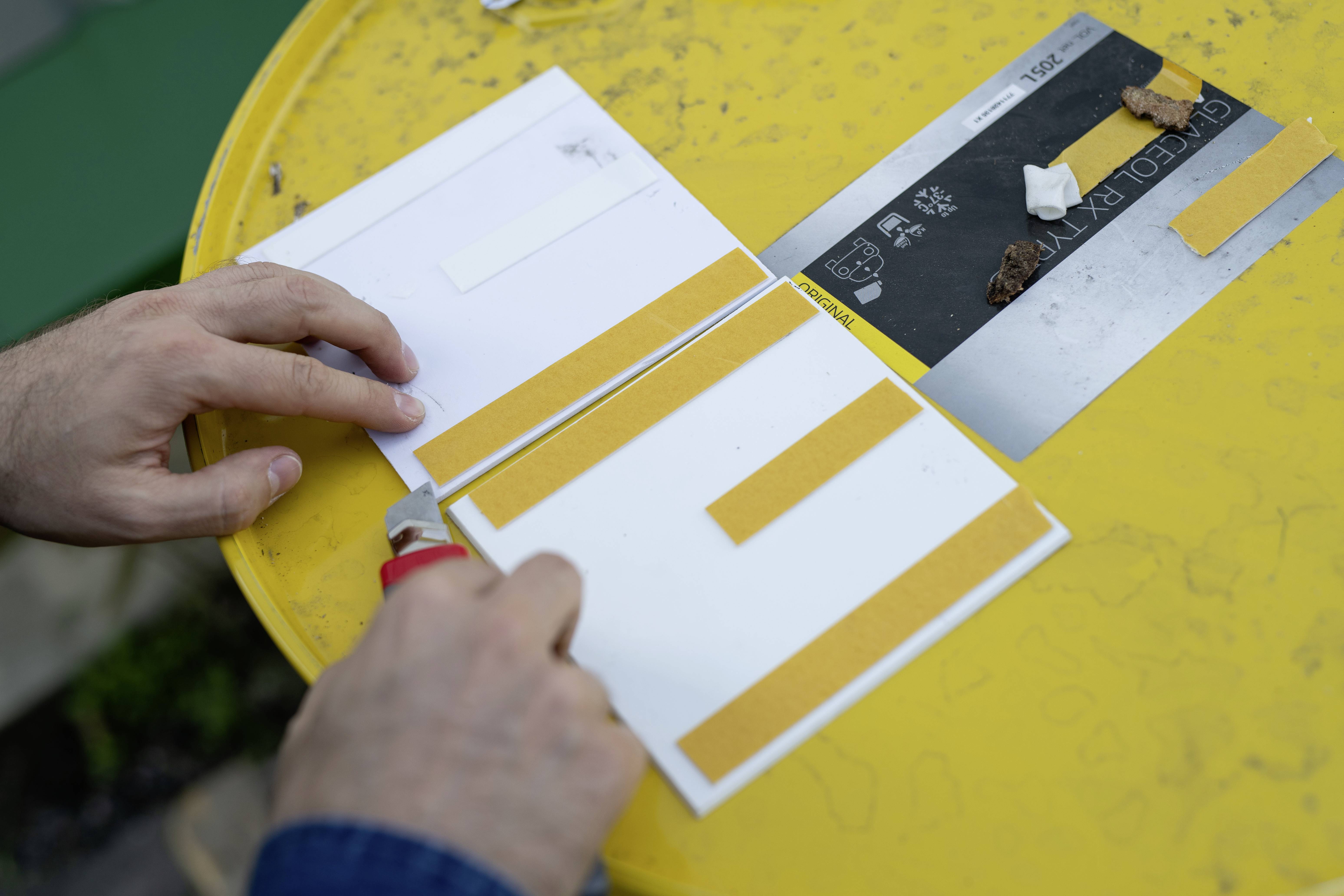 En person klipper gult tape på et gult bord for at sætte det på hvide kort. Et sort board med småting er ved siden af.