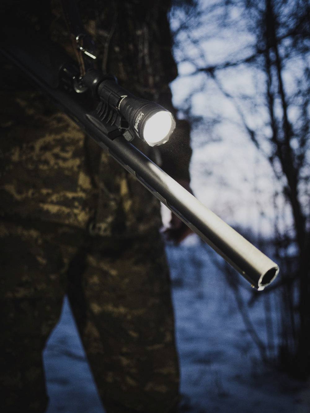 En person i camouflage holder et gevær med lommelygte i skoven ved tusmørke.