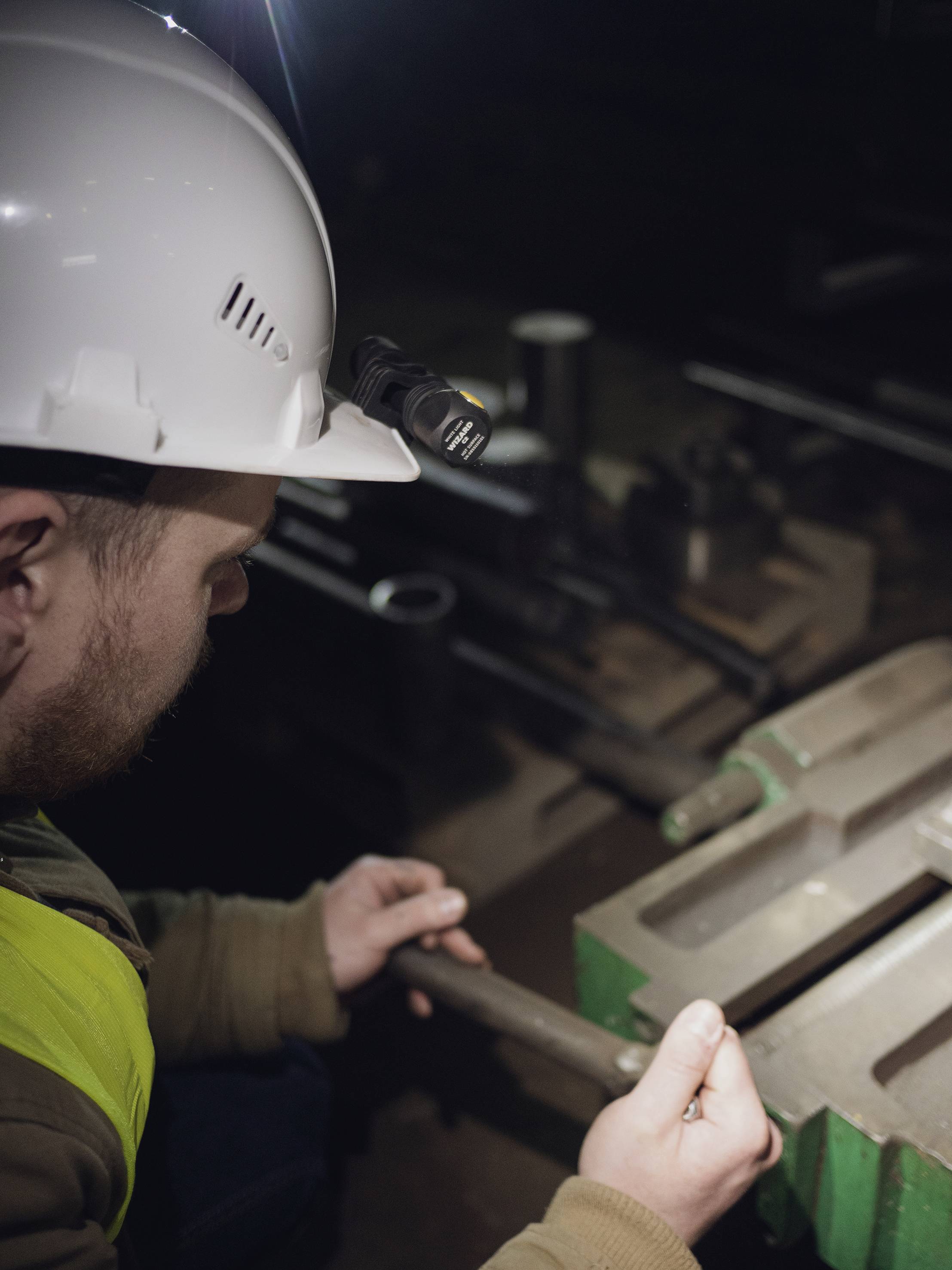 En arbejder med hjelm og pandelampe bearbejder et emne i et mørkt værksted, koncentreret om en præcis opgave.