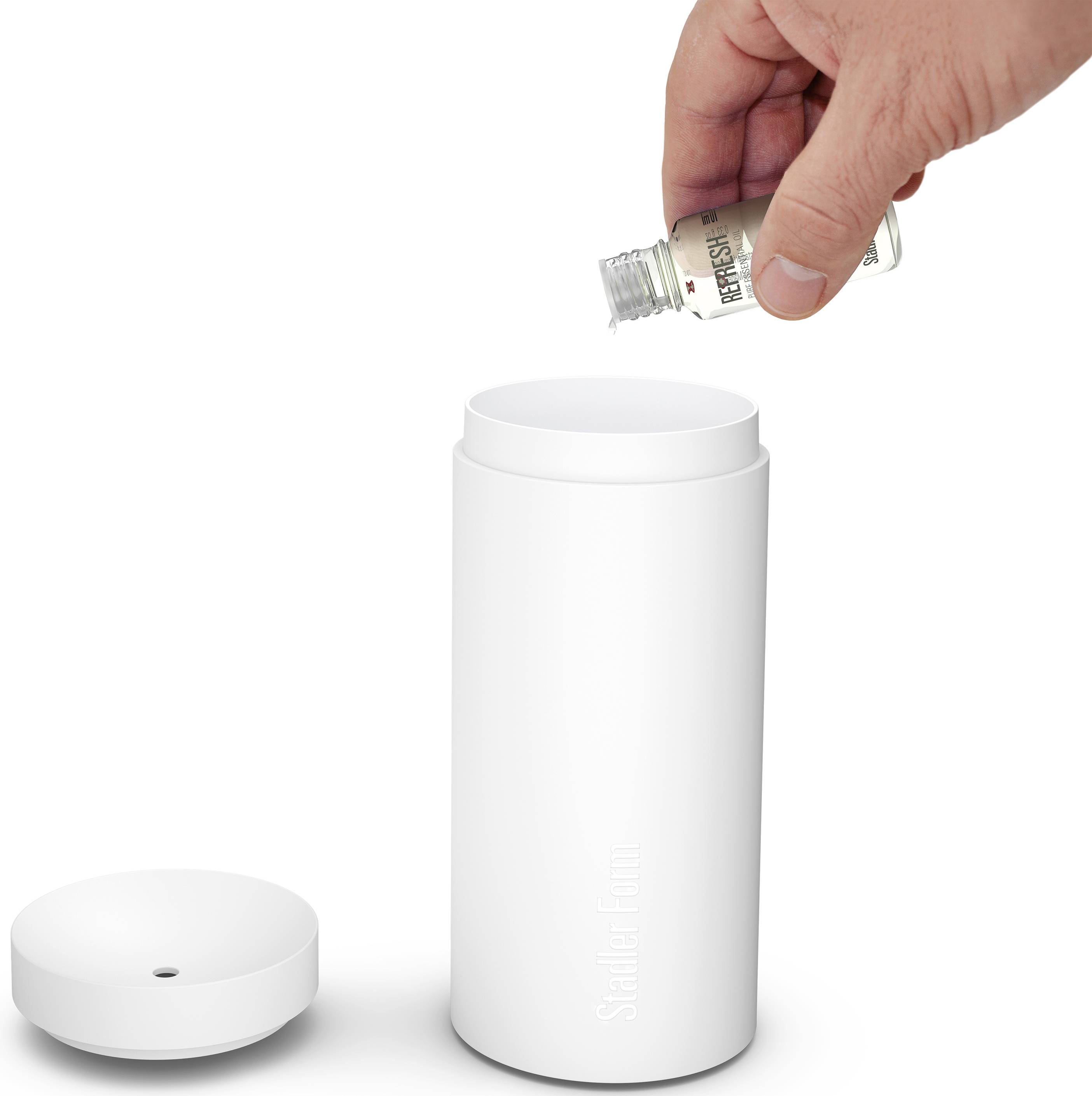 En hånd hælder æterisk olie fra en lille flaske i en hvid aroma-diffuser. Låget ligger ved siden af.