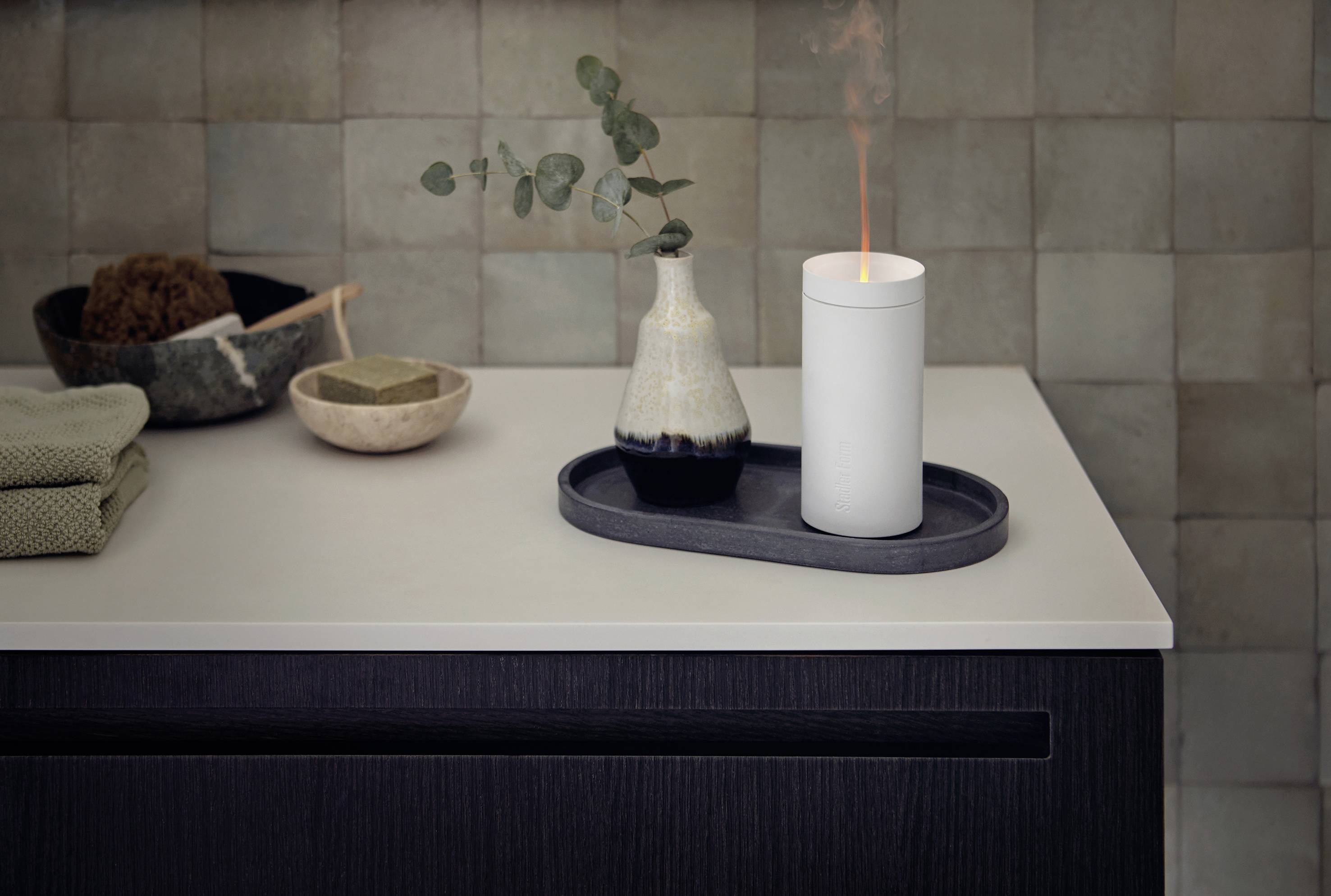 Et stearinlys brænder på et bakke ved siden af en vase med en eukalyptusgren. Begge står på en moderne bordplade i badeværelset.