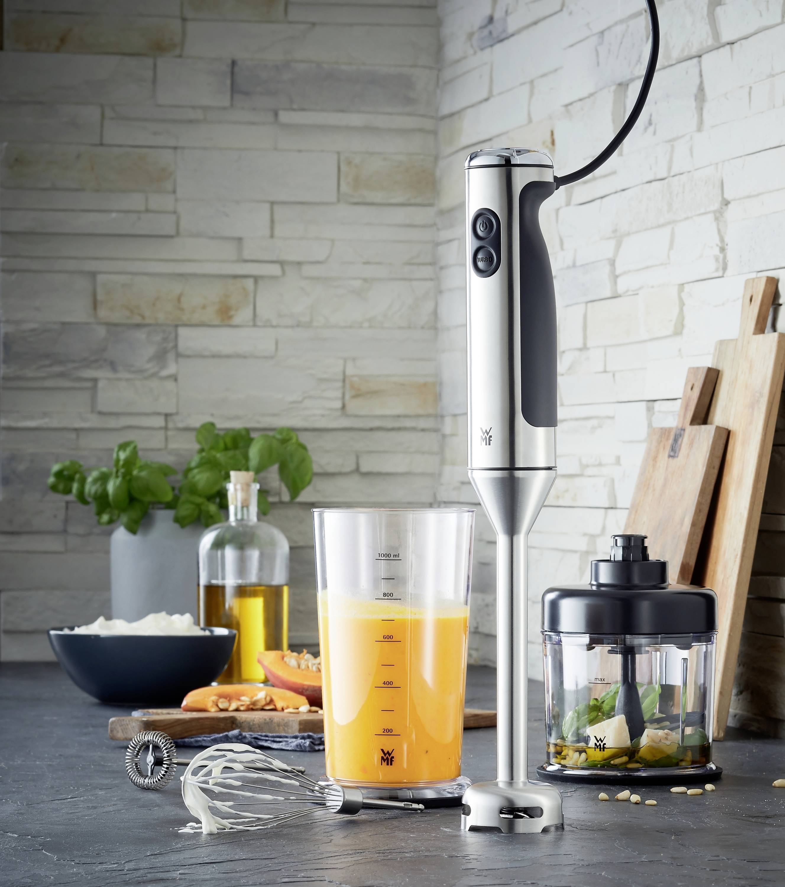 Et stavblendersæt på et køkkenbord, omgivet af et målebæger med appelsinjuice, ingredienser i en beholder og køkkenredskaber.