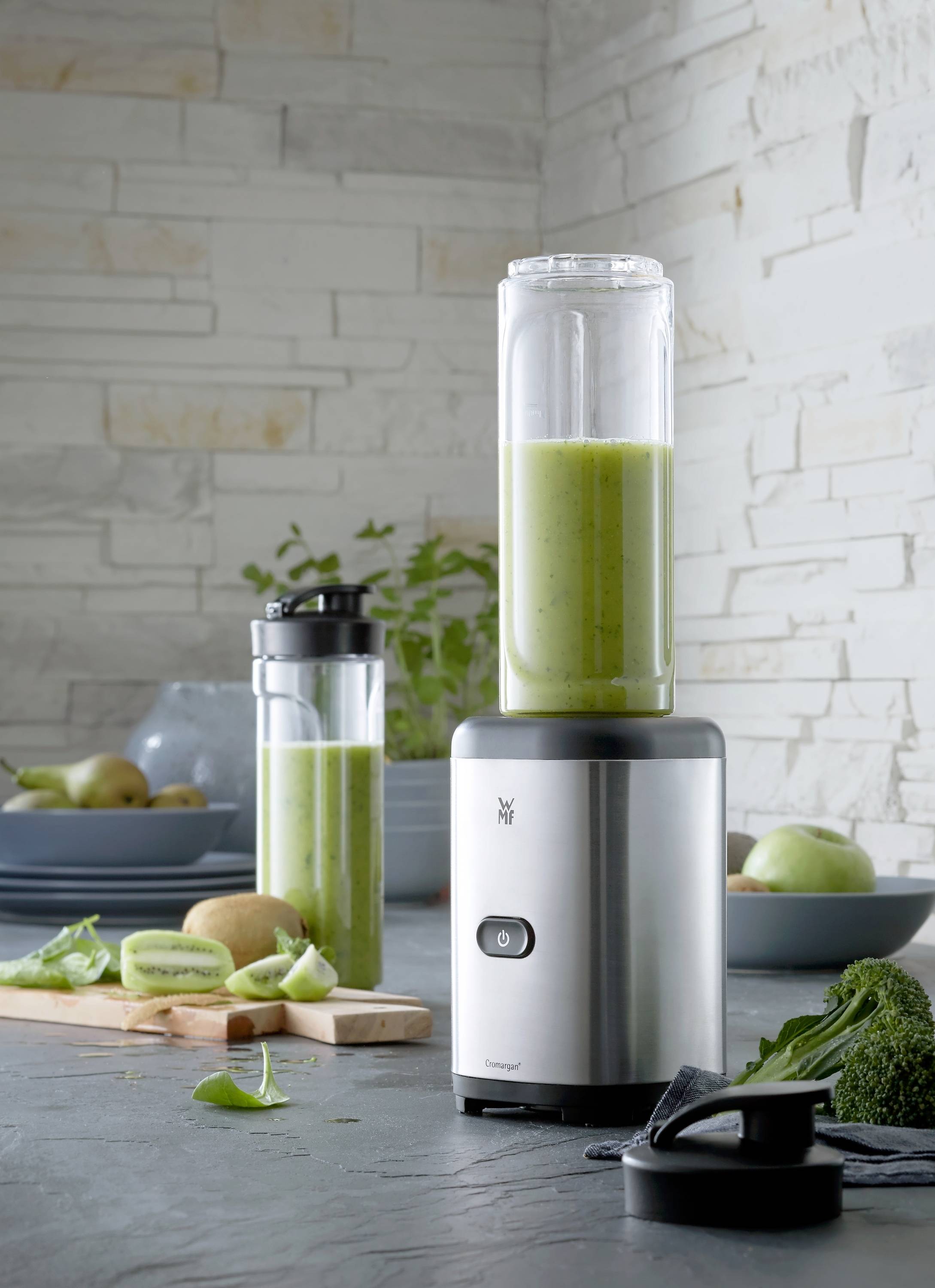 En sølvfarvet blender med et glas grøn smoothie står på et køkkenbord, omgivet af kiwi og spinat. I baggrunden et andet glas.