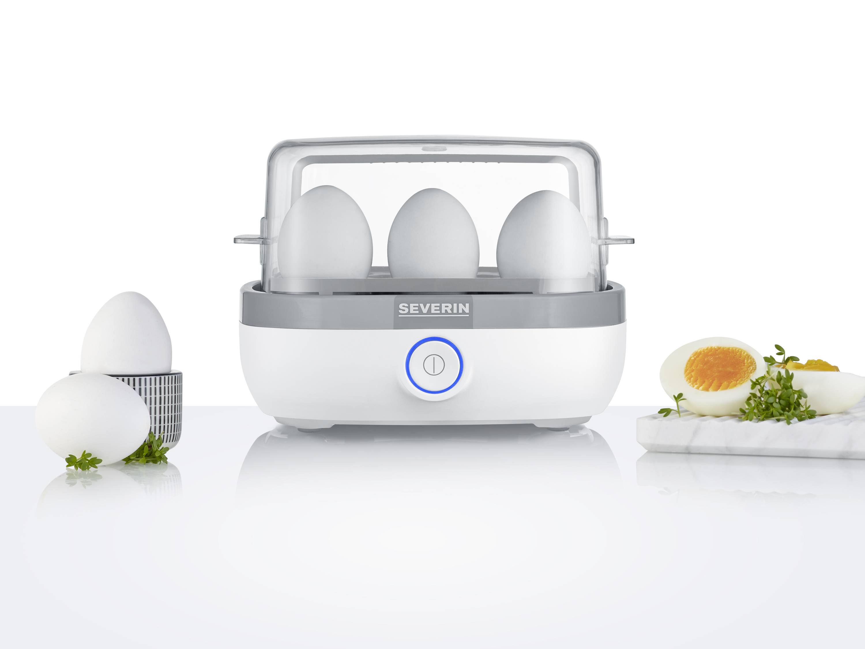 En æggekogermaskine med fire æg indeni og et kogt, halveret æg på et bræt. Æggekogeren er hvid med grålige accenter.