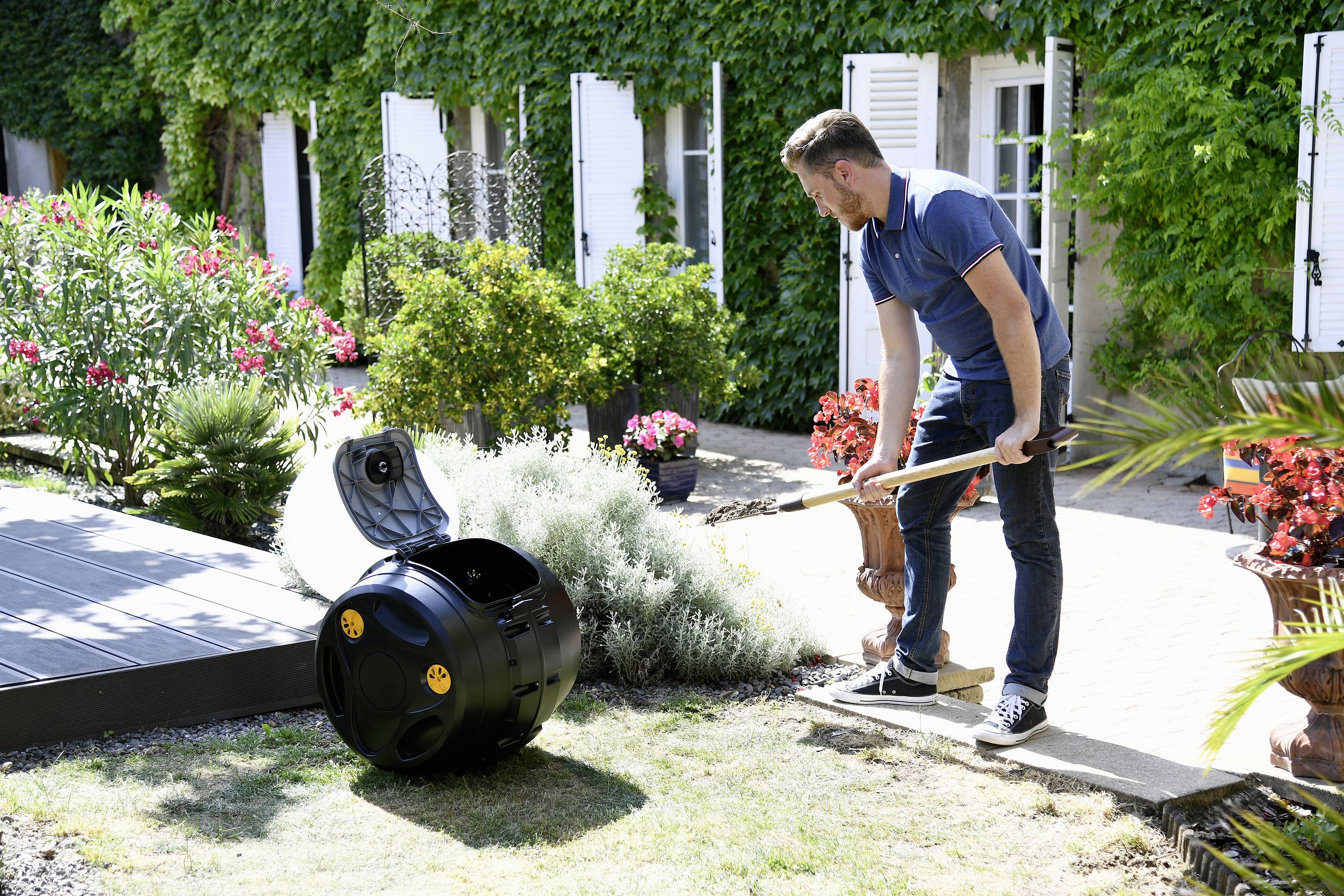 En mand bruger en kompostbeholder i haven. Han fylder beholderen med haveaffald for at fremstille kompost. I baggrunden ses planter.