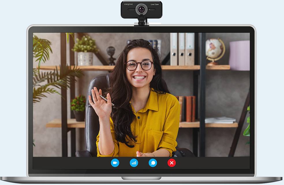 En kvinde sidder ved et skrivebord og smiler, mens hun vinker ind i webcammen. I baggrunden er der bogstakke synlige.