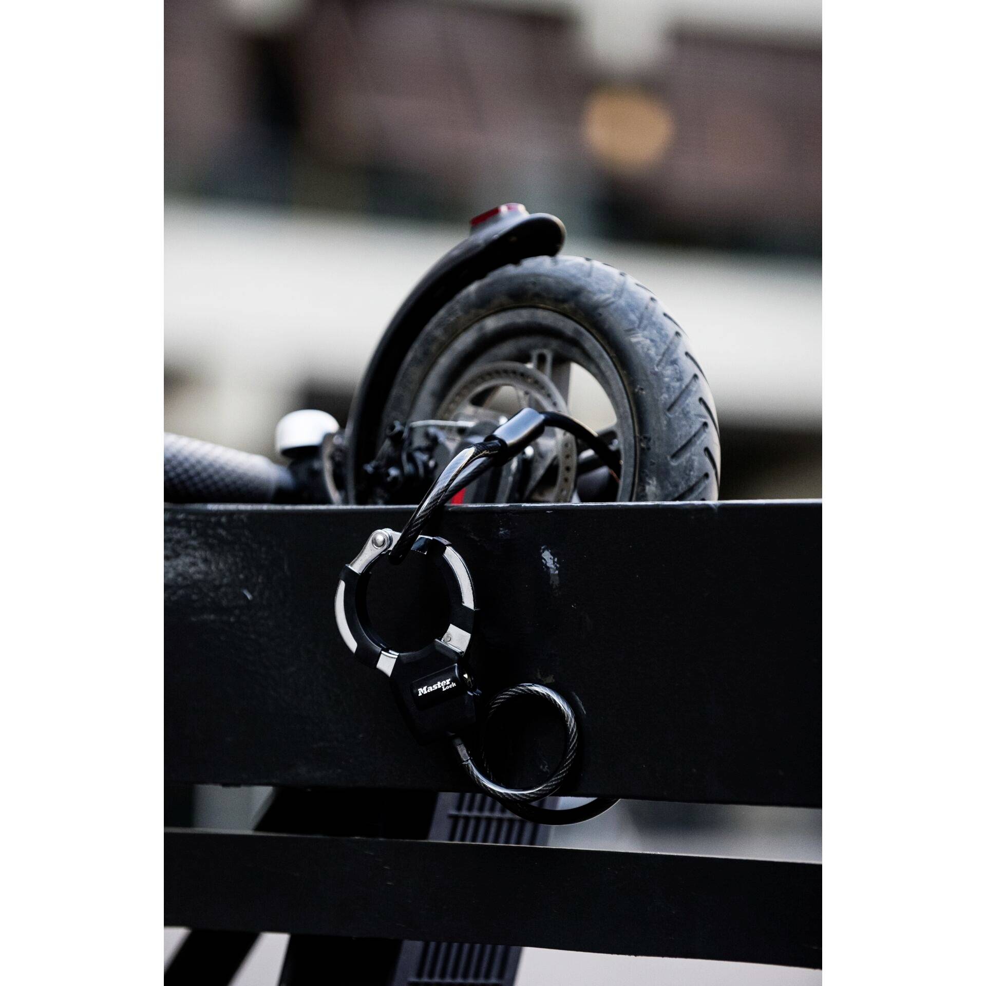 En elektrisk scooter er låst fast med en sort lås ved en metalgitter. Fokus er på baghjulet og låsen.