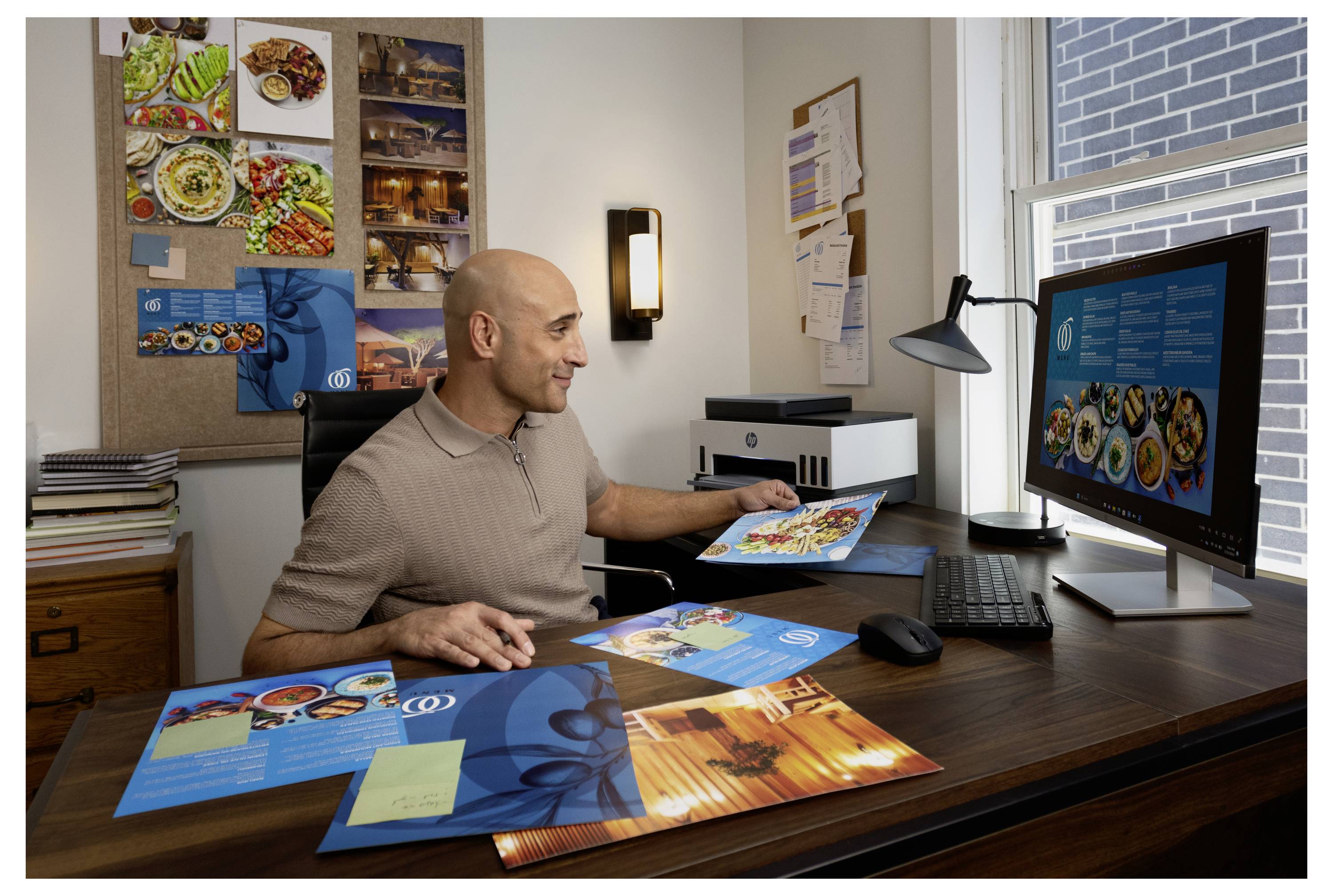 En mand gennemgår farverige menu- eller brochuredesigns ved et skrivebord med en computer, der viser layoutprøver på hjemmekontoret.
