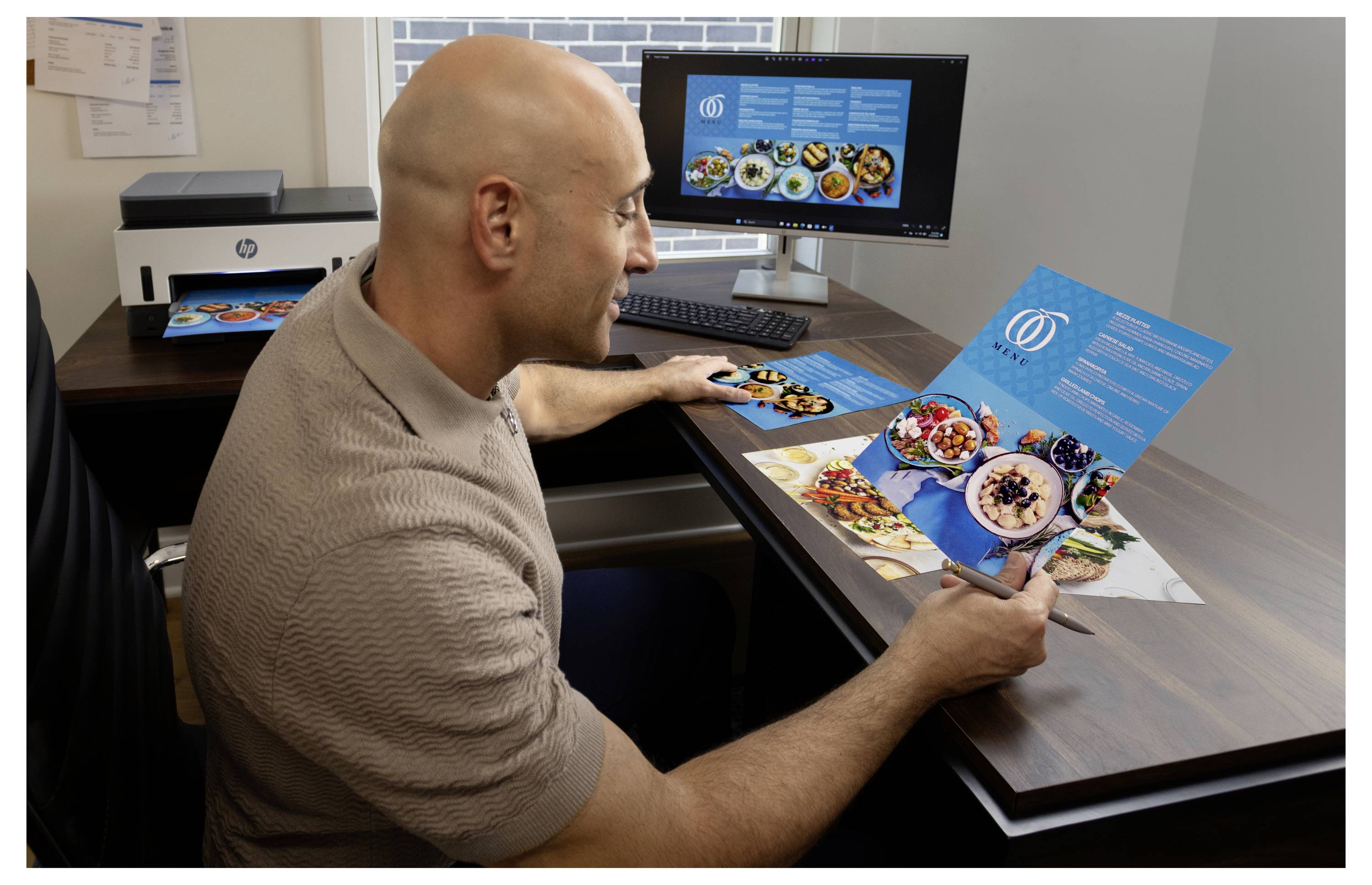 En mand anmeldelser et blåt madbrochureopslag på skrivebordet, hvor han sammenligner den trykte side med designet vist på hans computermonitor.