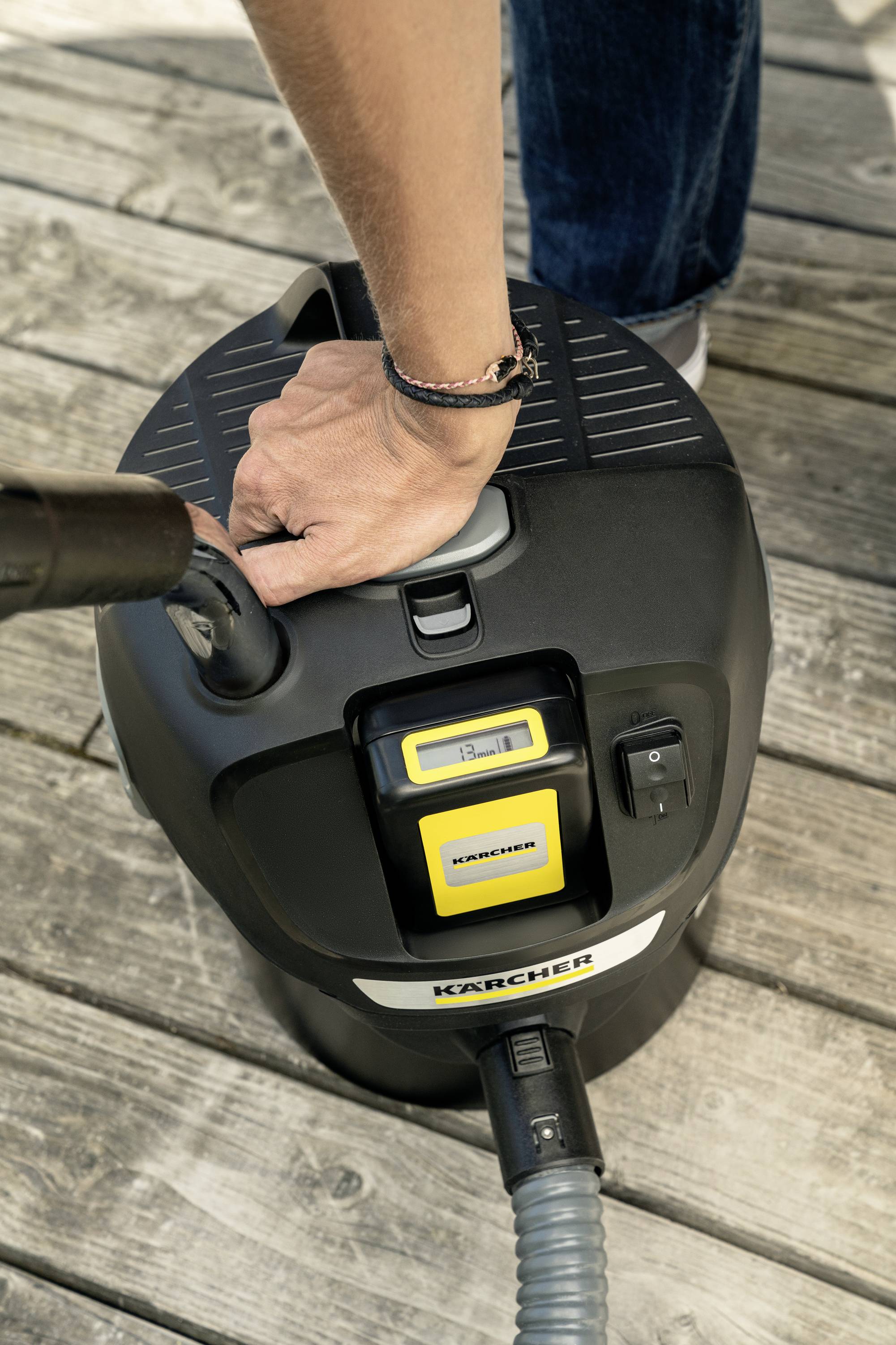 En person rengør en træterrasse med en våd-/tørstøvsuger. Hånden holder håndtaget på apparatet. Apparatet står på terrassen.